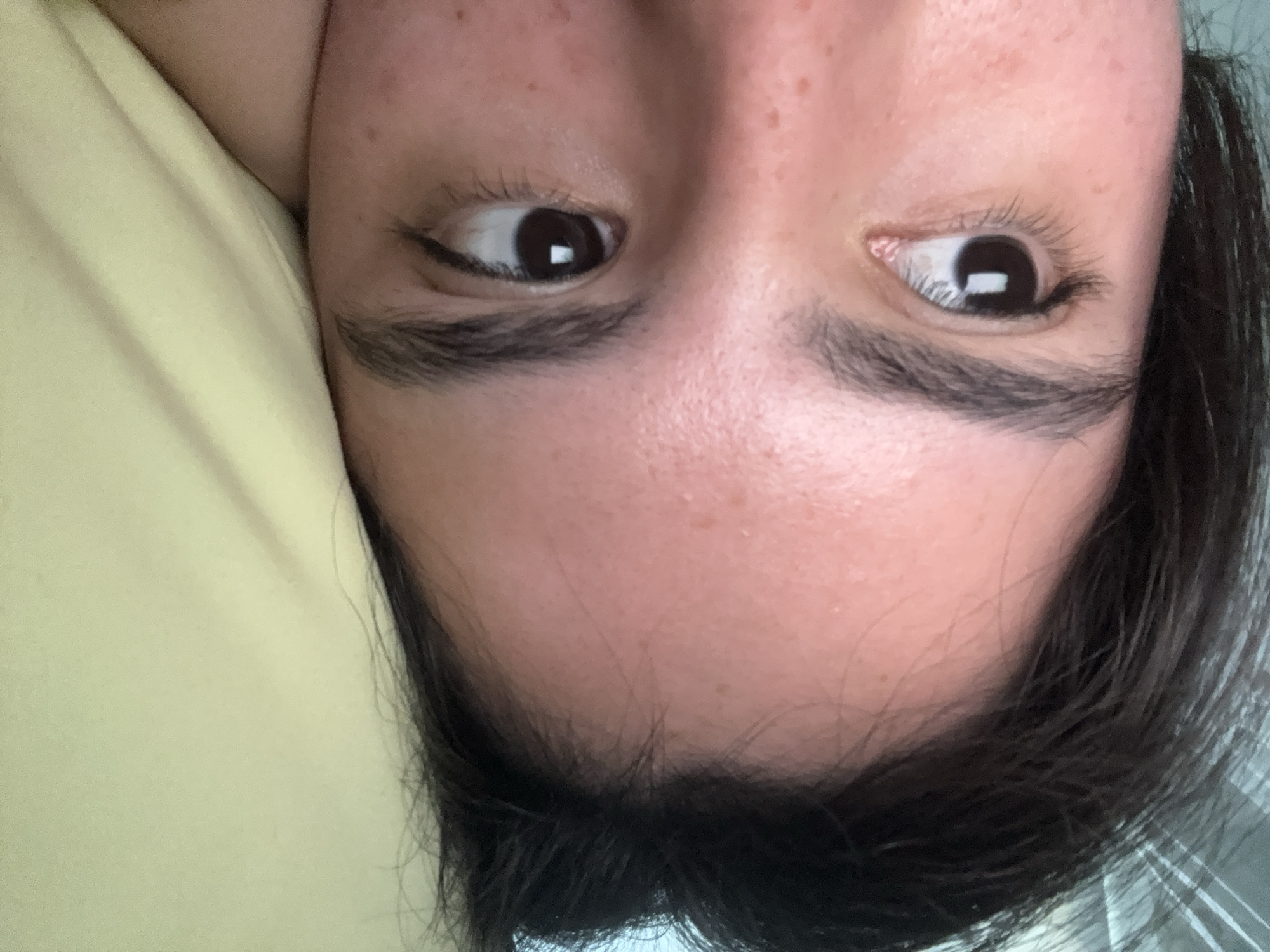 A close-up, tilted image shows the upper portion of a person's face. The person has brown eyes that are glancing to the side, dark eyebrows, and fair skin with some freckles. Dark hair is visible along the forehead and on the right side of the frame. The person is lying down on a yellow-green pillow or surface. The environment is undiscernible. The image's location cannot be determined from the image alone.