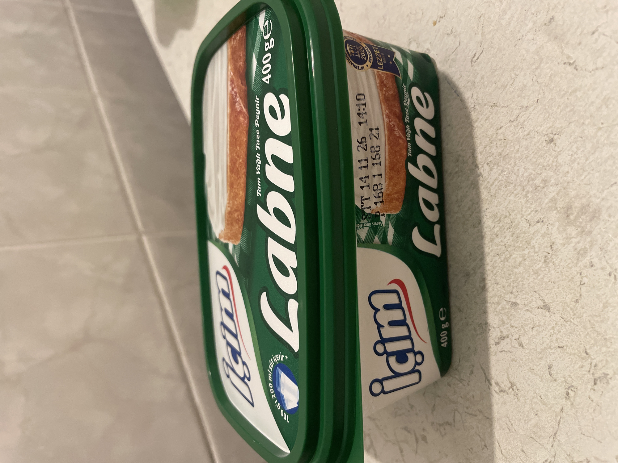 A close-up, eye-level, indoor shot shows a green plastic tub of "Labne" cheese. The tub is positioned on a light-colored countertop with a mottled texture, and a tiled wall forms the background. The tub's lid displays the brand name "İçim" in blue and red, along with the product name "Labne" in large white lettering. The text "Tam Yağlı Taze Peynir" (Full Fat Fresh Cheese) and "400 g e" are also visible on the lid. The tub itself has "İçim" written on it in a similar style, and "400 g e" is printed on the side. The lighting is overhead, creating subtle shadows. The scene appears to be a kitchen counter. The image does not provide any information about the city of Kadıköy, Turkey, or any people.