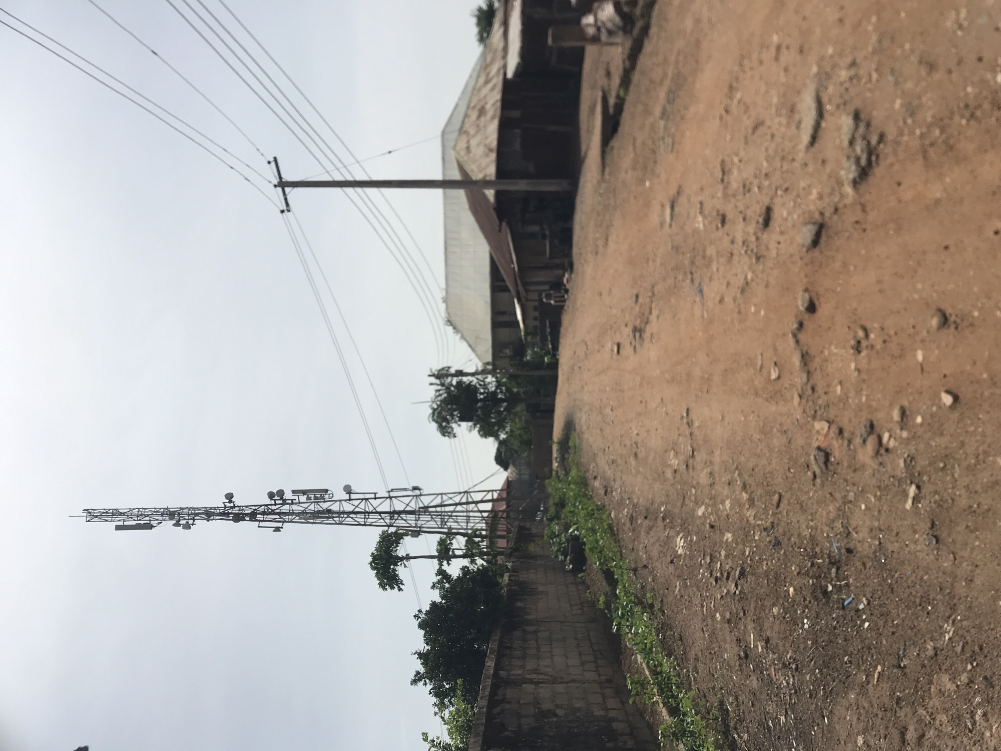 This image depicts a rural or semi-urban outdoor scene in Rafin Par, Nigeria, captured during the daytime under an overcast sky.
In the foreground, a dusty, unpaved dirt road or path occupies the right side of the frame, showing uneven terrain with loose earth, small stones, and scattered debris. Parallel to this path on the left is a low concrete block wall, partially overgrown with green grass and small shrubs.
Dominating the midground on the left is a tall, metal lattice telecommunications tower, equipped with multiple antennas and satellite dishes, standing against the bright sky. Above and to the left of the tower, several electrical or telephone lines stretch horizontally across the sky, supported by a utility pole partially visible on the far left.
In the background, behind the wall and tower, several single-story buildings with corrugated metal roofs are visible, suggesting homes or small businesses. Green trees and dense foliage are scattered throughout the background and around the base of the tower.
The sky is uniformly bright and light gray, indicating an overcast day with no direct sunlight. No people or specific activities are discernible in the image. The scene conveys a functional environment with essential infrastructure. There is no visible text.