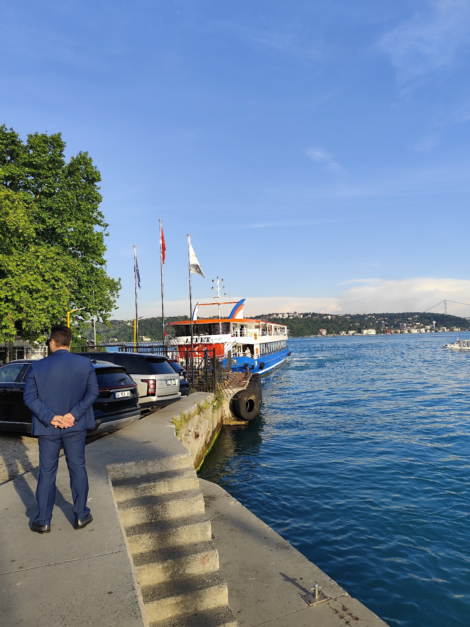 The image presents a vibrant waterfront scene in Sarıyer, Turkey, likely along the Bosphorus Strait, bathed in the late afternoon or early evening sun. The clear blue sky is adorned with only a few wispy clouds.
In the foreground, a man dressed in a dark blue suit stands with his back to the viewer, hands clasped behind him, seemingly observing the activity on the water. To his immediate left, several cars are parked on a paved area, including a dark SUV/sedan displaying the license plate "34 MCK 60" and a lighter-colored SUV, possibly a Range Rover, with a partially visible plate "34 CL 112". A large, green-leafed tree is prominent on the left, casting a shadow.
Adjacent to the parked vehicles, a concrete pier features a set of stone steps descending towards the water. Three flags fly from poles: a red flag (suggesting the Turkish national flag), a blue flag, and a white flag. A blue and white ferry, adorned with orange trim and the name "AFFET" visible on its side, is either docked or preparing to depart near the pier. Black rubber fenders are attached to the pier's edge.
The deep blue water exhibits gentle ripples. Across the strait, a lush, hilly coastline is visible, densely populated with buildings. In the distance to the right, a large suspension bridge, characteristic of the Bosphorus, spans the water. Another smaller, white boat can be seen further out on the water. The overall impression is one of a busy yet scenic urban coastal environment.