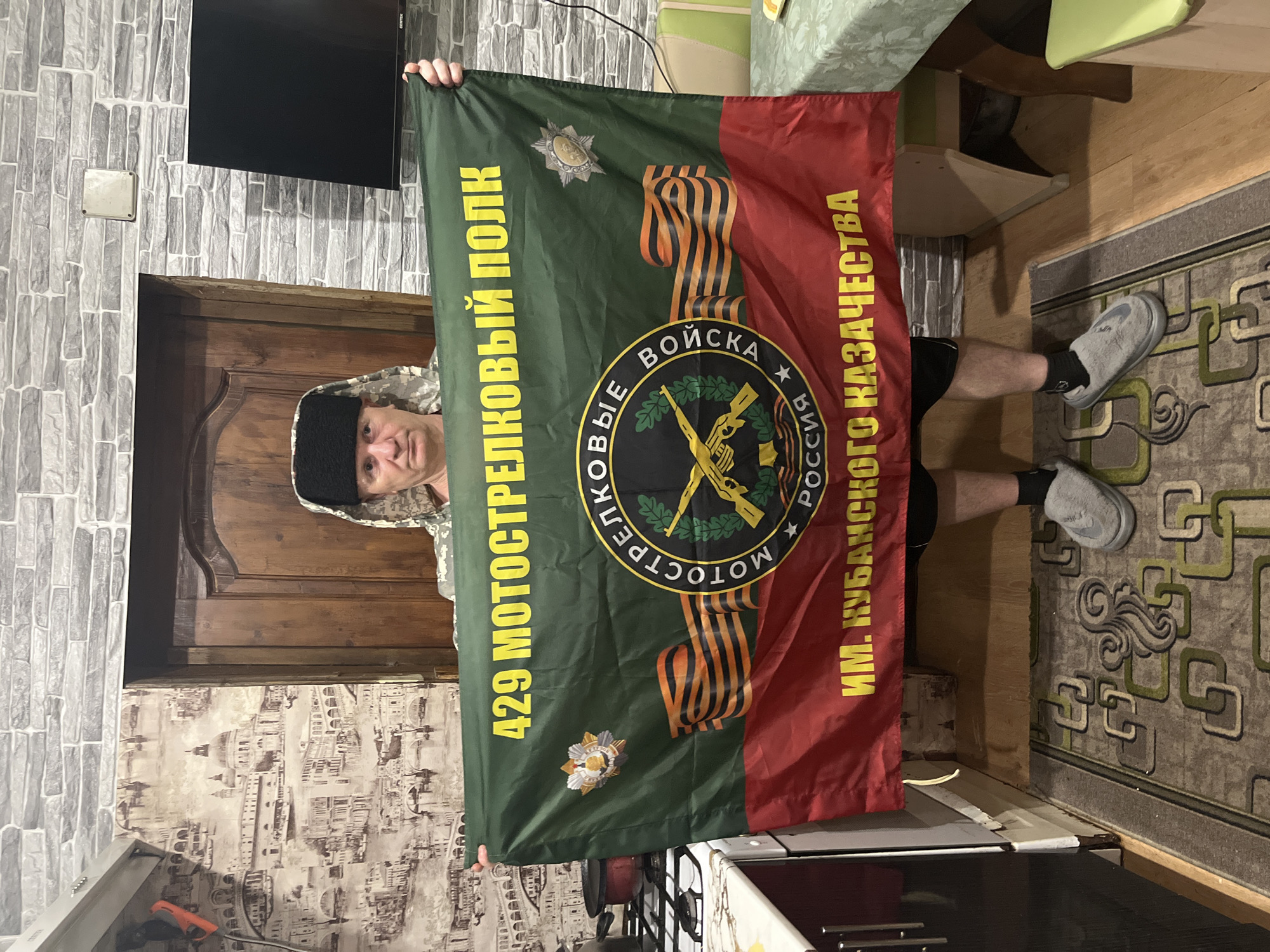 A man stands indoors, holding a flag. The flag is divided vertically into green and red sections, with the text "429 мотострелковый полк" on the green side and "ИМ. Кубанского казачества" on the red side, both written in yellow. The flag also features the ribbon of Saint George and a logo that contains crossed rifles. The man is wearing a camouflage-patterned jacket and a black hat. He appears to be in a room, with a wooden door behind him and patterned wallpaper. The location is Vesyoloye, Russia.
