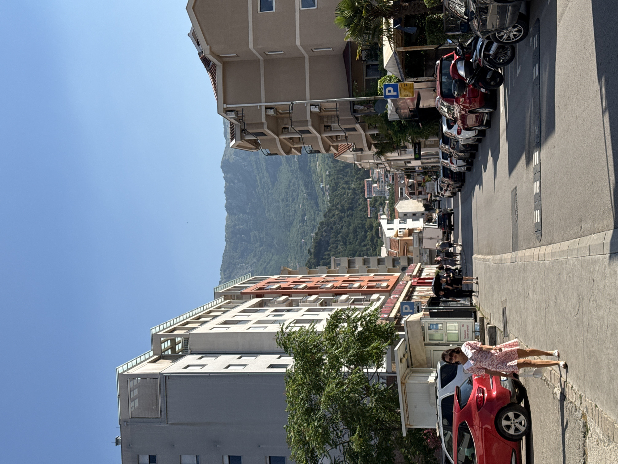 A medium shot captures a street scene in Budva, Montenegro, on a clear, sunny day. The perspective is looking down a street lined with multi-story buildings and parked cars. In the foreground, a woman in a pink floral dress walks on the sidewalk, holding a white shopping bag. A red car is parked to her left. The street is filled with parked cars on both sides. Buildings on the left are modern, with balconies and windows. The building on the far left is a light gray structure with a contrasting orange section. The buildings on the right are a sandy beige color, also featuring balconies and air conditioning units. In the background, lush green mountains rise under a clear blue sky, with some smaller buildings visible on the hillsides. A parking sign with a blue "P" is visible on the right side of the street. The overall impression is a typical urban environment with a natural backdrop.