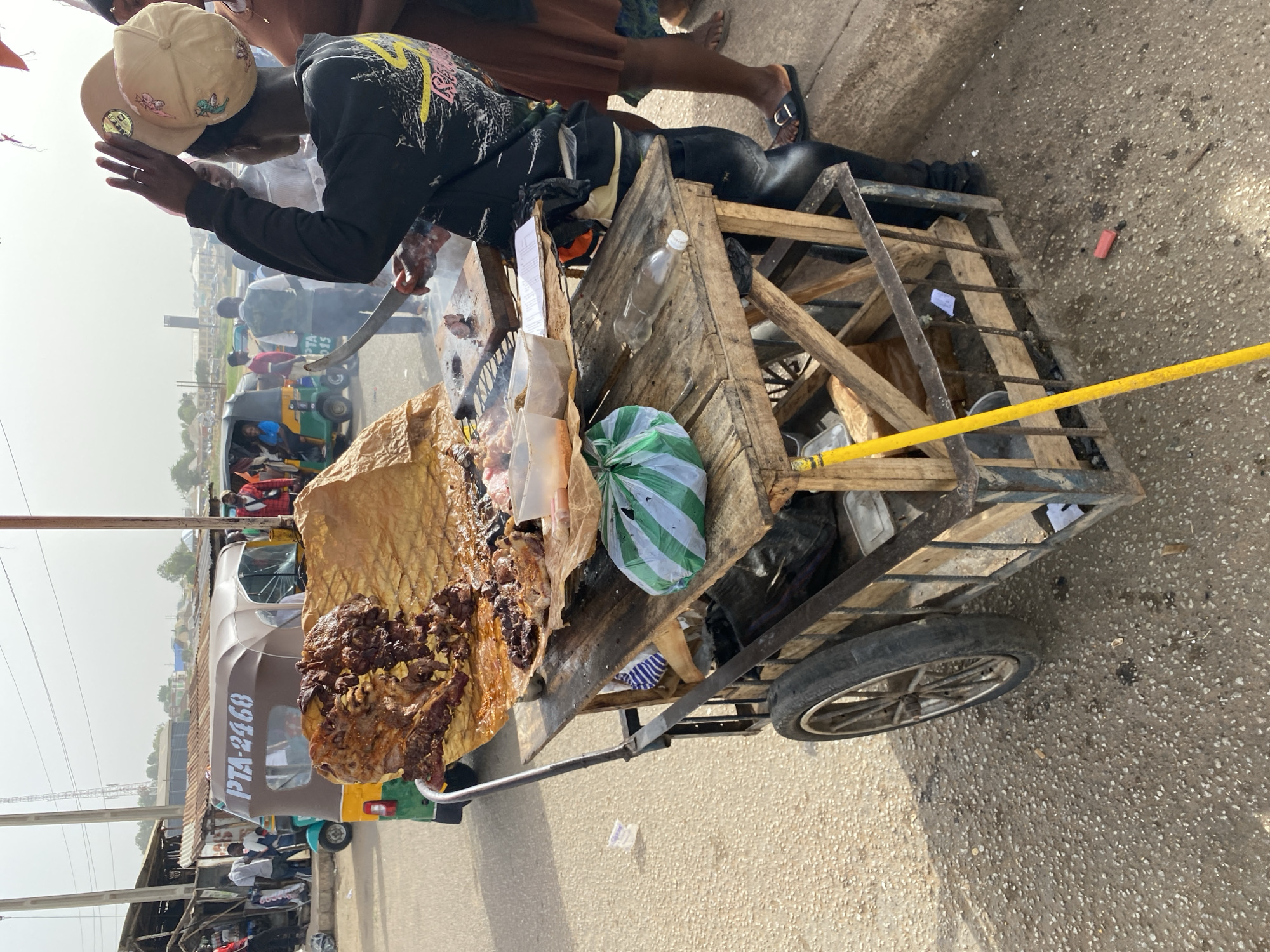 The image depicts a street food vendor in Bukuru, Nigeria, preparing and serving what appears to be roasted meat. The vendor, wearing a hat and long-sleeved shirt, is cutting the meat with a large knife. The meat is displayed on a wooden cart with two wheels, along with other items such as plastic bags and a water bottle. In the background, there is a three-wheeled vehicle (tuk-tuk) and other people. The setting is a busy street.