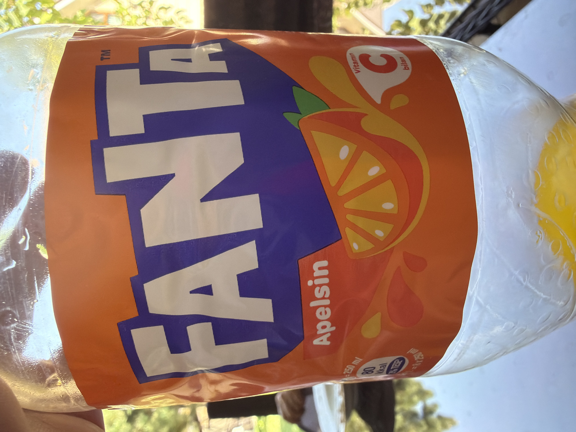 The image shows a close-up of a Fanta bottle with an orange label. The bottle is held by a hand, and the background is blurred, showing some greenery and sunlight. On the label, the Fanta logo is prominently displayed in white letters against a purple background. Below the logo, "Apelsin" is written in white text. There are also graphics of an orange slice and splashes of orange liquid. A small section on the right side of the label shows "Vitamin C" and "bilan". On the left side, there are nutritional facts indicating "250 ml", "80 kkal", and a symbol with a fork and knife. The bottom of the bottle is clear plastic, and some yellow liquid is visible inside. The scene is outdoors, with natural light. The location is Koshkurgon, Uzbekistan.