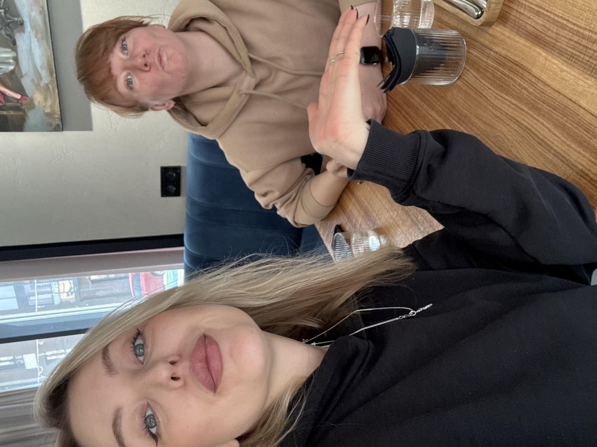 Two women sit at a wooden table in an indoor setting. One woman, with blonde hair and wearing a black hoodie, is in the foreground. The other woman, with reddish-brown hair and wearing a tan hoodie, is partially visible in the background. The table holds glasses and a black object. A window in the background suggests an urban setting. The location may be in Krasnoyarsk, Russia.