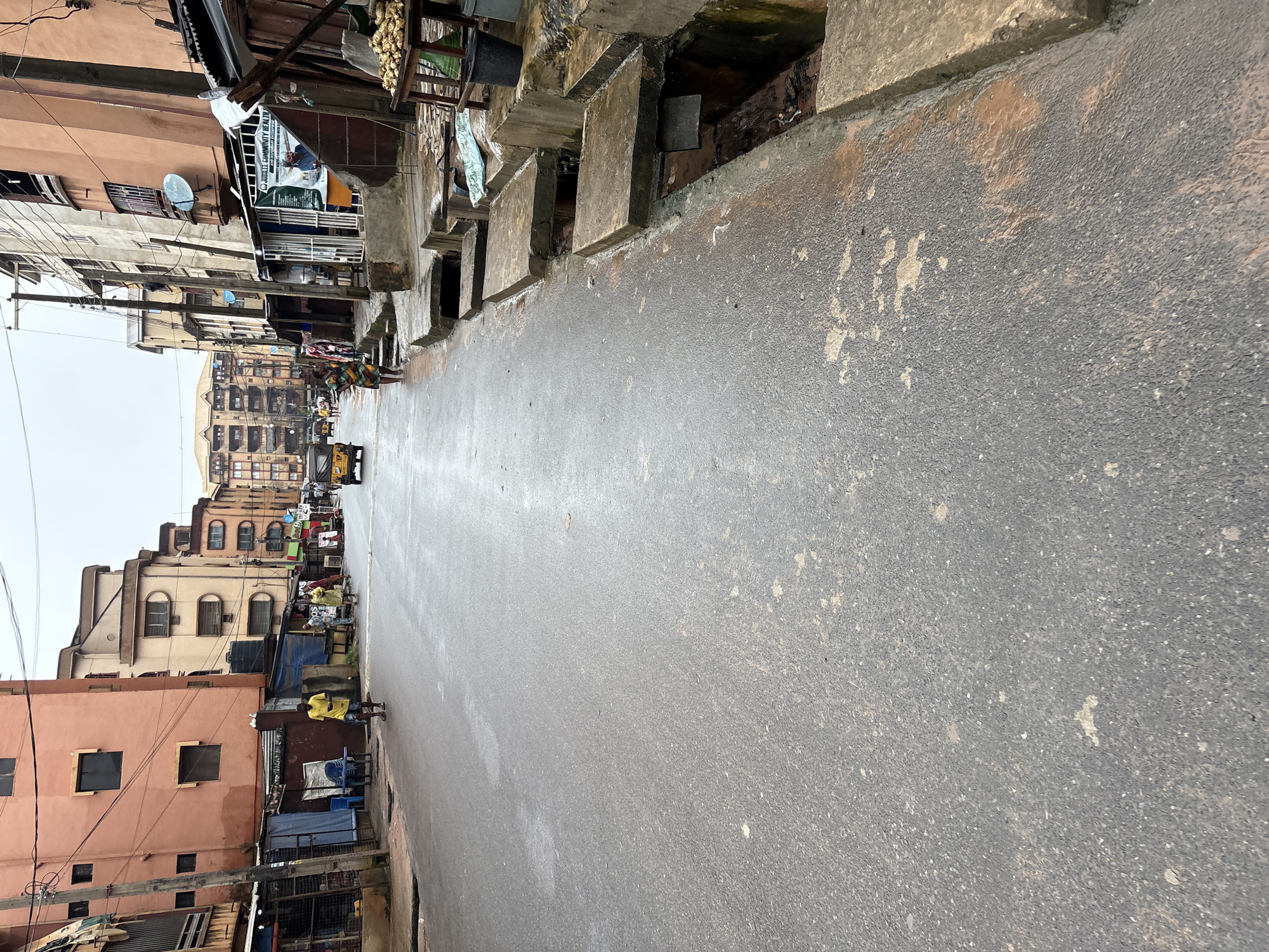 This is a street-level, medium-distance, eye-level shot of a street in Onitsha, Nigeria. The scene depicts a moderately busy street with buildings lining both sides. The road surface is paved with asphalt and appears wet, possibly from recent rain, with some patches of discoloration and debris.
On the left side of the street, there are multi-story buildings with visible windows and balconies. Some of these buildings have shops or stalls at ground level, with tarpaulin coverings and various items displayed. People are seen walking on the sidewalk and in the street. One man in a yellow t-shirt and blue shorts is walking towards the camera, while others are further down the street.
On the right side of the street, there are more multi-story buildings and a more pronounced drainage system with concrete slabs covering open channels. A small yellow motorized rickshaw or "Keke Napep" is visible in the distance, along with other pedestrians. A vendor's stall with produce, possibly yams, is set up on the right side. A black bucket is placed near the stall.
The sky is overcast, suggesting a cloudy or possibly rainy day. The overall impression is a typical urban street scene in a Nigerian city, characterized by its vibrant architecture, street life, and the presence of commercial activities.