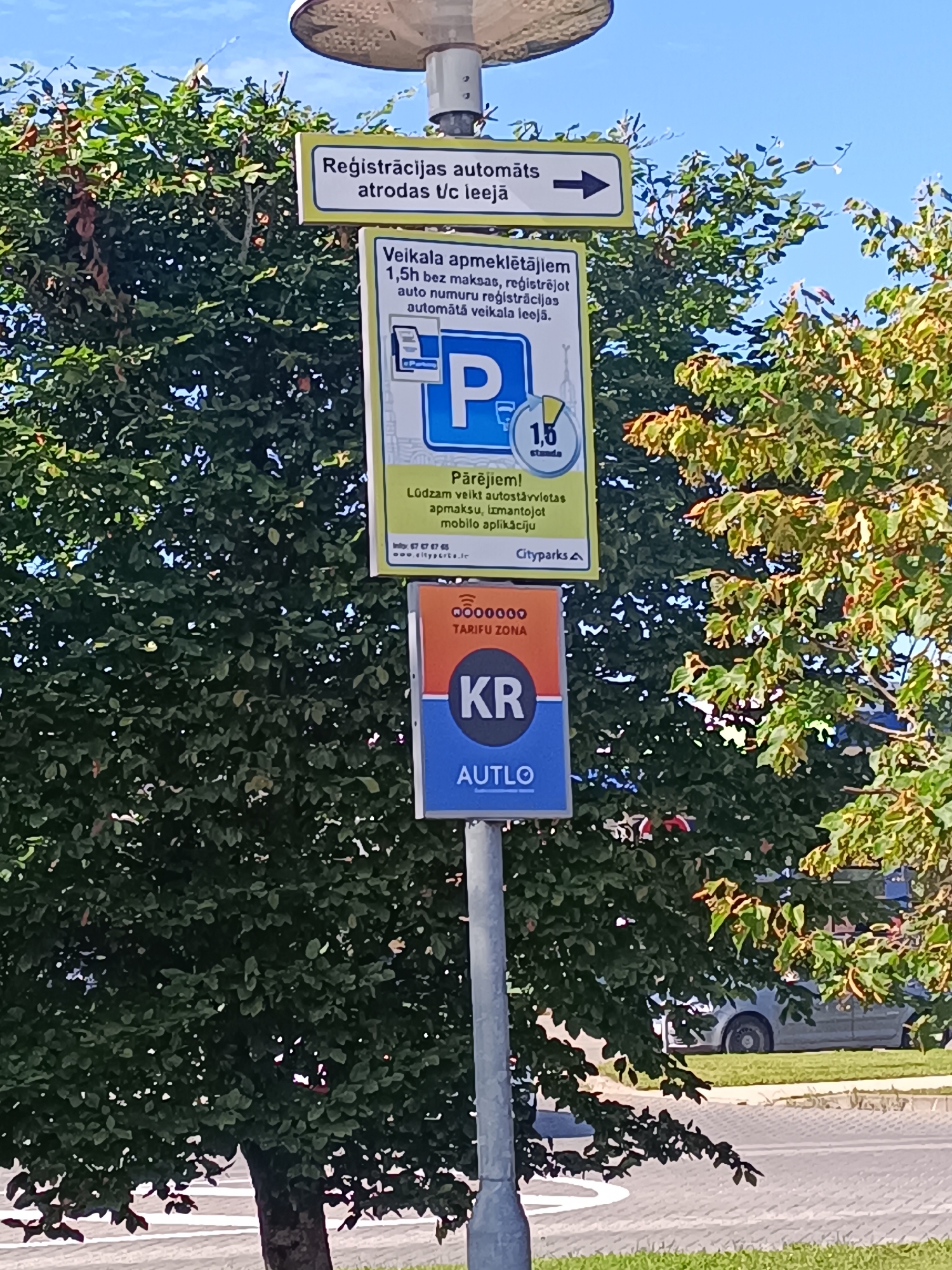 The image shows a pole with several signs in an outdoor setting, likely a parking area. The first sign, with a yellow border, reads "Reģistrācijas automāts atrodas U/c ieeja" and has an arrow pointing to the right. Below this is a larger sign with a yellow background and a blue "P" logo, indicating parking. Text on this sign reads "Veikala apmeklētājiem 1,5h bez maksas, reģistrējot auto numuru reģistrācijas automātā veikala ieeja." and "Pārējiem! Lūdzam veikt autostāvvietas apmaksu, izmantojot mobilo aplikāciju." Below this, a website and phone number are listed, along with "Cityparks A". The bottom sign is orange and blue, with "000000 TARIFU ZONA KR AUTLO" prominently displayed. The background is dominated by lush green trees, suggesting a well-maintained area. The sky is visible above, and a car is partially seen in the background on the right. The overall scene appears to be in a public space, possibly near a shopping center or commercial area, given the references to a shop and parking fees. The presence of Latvian text indicates the location is in Latvia.