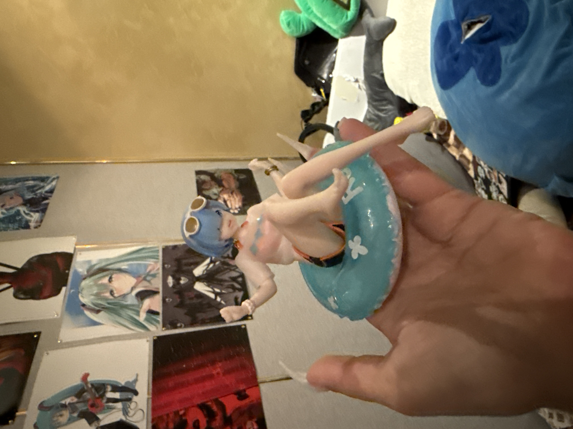 In Odesa, Ukraine, a close-up shot focuses on a hand holding a figurine of a female character with blue hair, wearing sunglasses and a swimsuit, seated on a blue inflatable ring. The figurine appears to be a collectible or toy. In the background, several anime-style posters are visible on a wall, depicting various characters. One poster shows a character with green hair and pigtails, another with a black and white design. Further in the background, other objects and plush toys are partially visible, including a green plush toy and a large blue plush item with a floral design. The lighting suggests an indoor environment, possibly a room or display area.