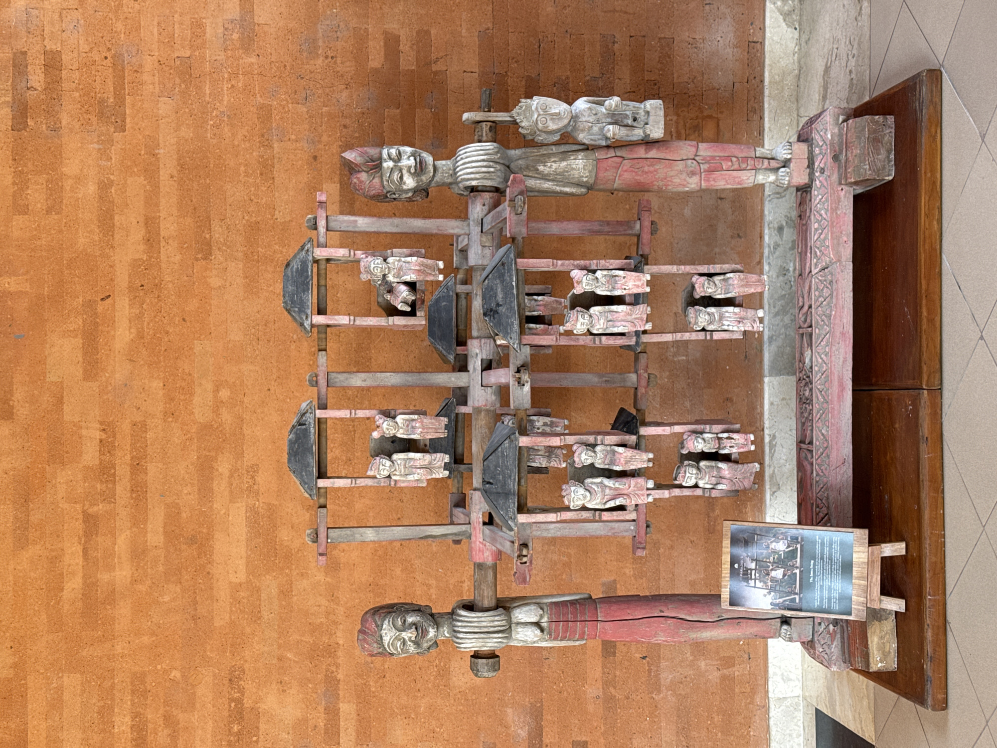 The image shows a wooden sculpture on display, photographed from above, at the Garuda Wisnu Kencana Cultural Park in Kuta Selatan, Indonesia. The sculpture consists of two larger carved human figures positioned horizontally at the top and bottom, connected by a series of shelves. Each shelf supports several smaller, carved human figures. The shelves are linked by vertical wooden supports, and black, spade-shaped components are attached. A sign with text and an image sits at the lower right. The sculpture is positioned next to a corner, with a wooden structure visible in the corner. The sculpture sits on a surface with rectangular tan blocks.