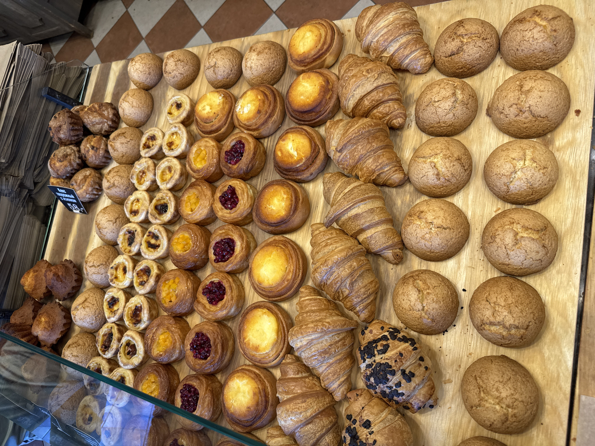 The image displays a variety of baked goods arranged on a wooden surface. The subjects are primarily pastries and bread, including croissants, buns, and filled pastries with fruit or cream. These items are neatly organized in rows, suggesting a display in a bakery or market.
The activity depicted is the presentation of food for sale. The lighting suggests an indoor setting, possibly during daylight hours due to the brightness of the scene. There are no people visible in the image.
Notable details include the diverse textures and colors of the baked goods, ranging from golden-brown croissants to cream-filled pastries with dark fruit toppings. The wooden surface shows grain patterns and slight wear, indicating a rustic or traditional setting. In the lower left corner, a price tag is partially visible with Cyrillic text. The text reads "25 58".