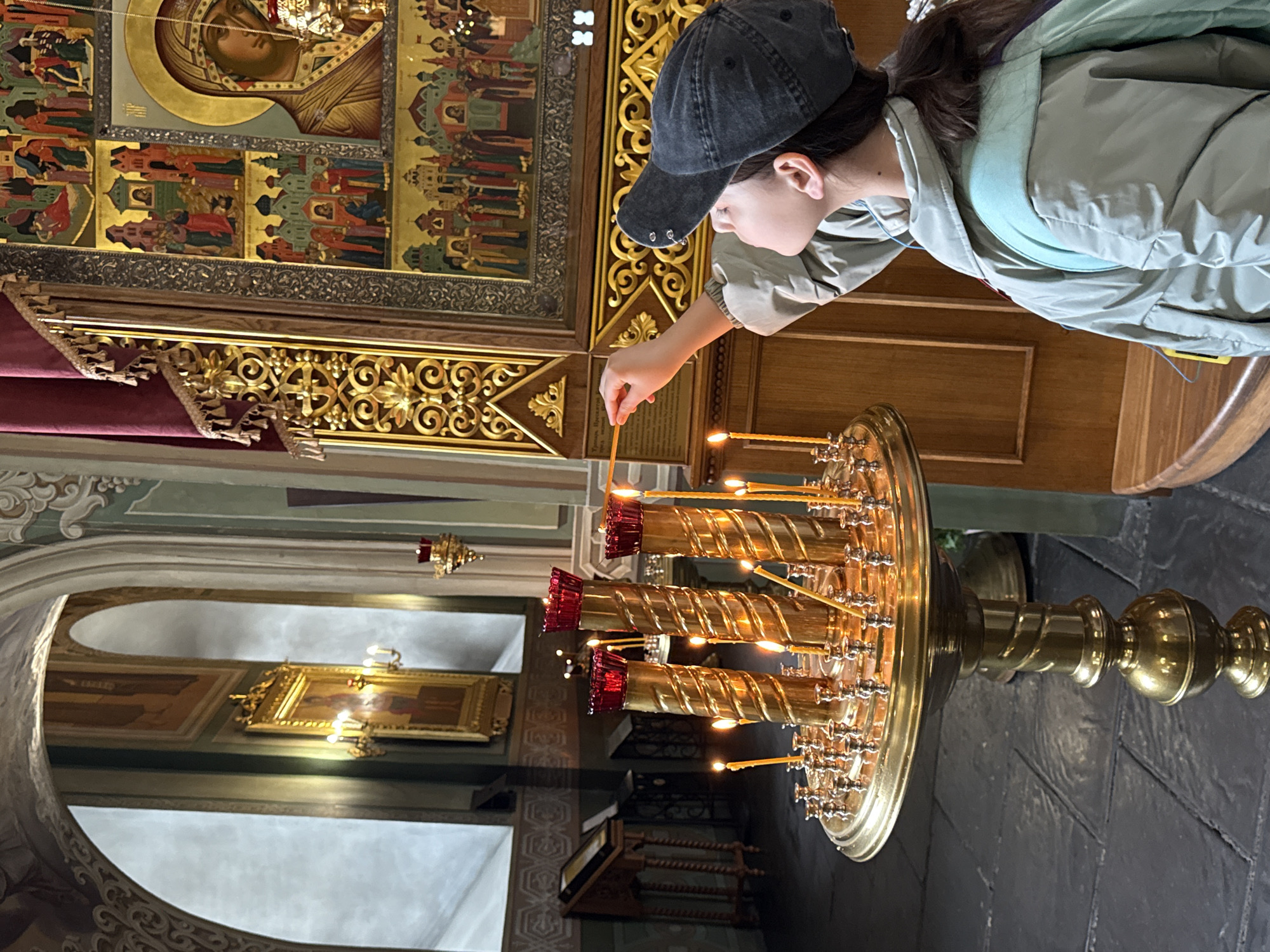 A young woman in a cap is lighting a candle in what appears to be an Orthodox church. The church's interior is ornately decorated with golden icons and carvings, characteristic of Russian Orthodox architecture. The woman is positioned near a large, multi-tiered candle stand made of brass or gold, with numerous lit candles. A window or archway in the background reveals another part of the church interior, with additional icons visible on the walls. Given the architectural style and the presence of Orthodox religious imagery, it is likely that this photograph was taken at Kazan Cathedral in Kazan, Russia.
