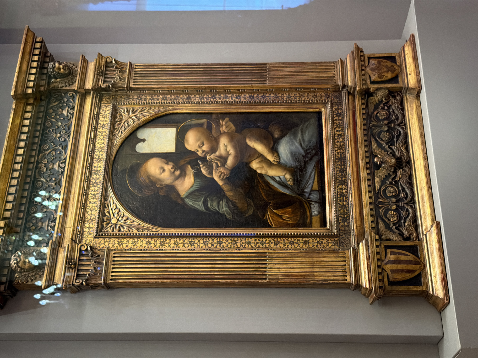 The image shows a painting of the Virgin Mary and the Christ Child in a richly ornate golden frame. The painting itself depicts a tender moment between mother and child, with Mary looking lovingly at Jesus, who is being held in her arms. The background of the painting is dark and somber, contrasting with the vibrant colors of the figures. The frame is elaborate, featuring carved details of foliage, mythical creatures, and heraldic shields. The painting is displayed on a wall in what appears to be a museum or gallery, with a gray wall and a white ledge beneath it. Reflections of light are visible on the surface of the painting, suggesting it might be behind glass, possibly for protection. The overall impression is one of historical and artistic significance, with the intricate frame drawing attention to the preciousness of the artwork. The style of the painting suggests it is an Old Master, likely from the Renaissance or Baroque period. The specific details of the frame, such as the shields and possibly family crests, could offer clues to the painting's provenance or the patron who commissioned it. The scene is static and devotional, intended to evoke a sense of reverence and spiritual contemplation.