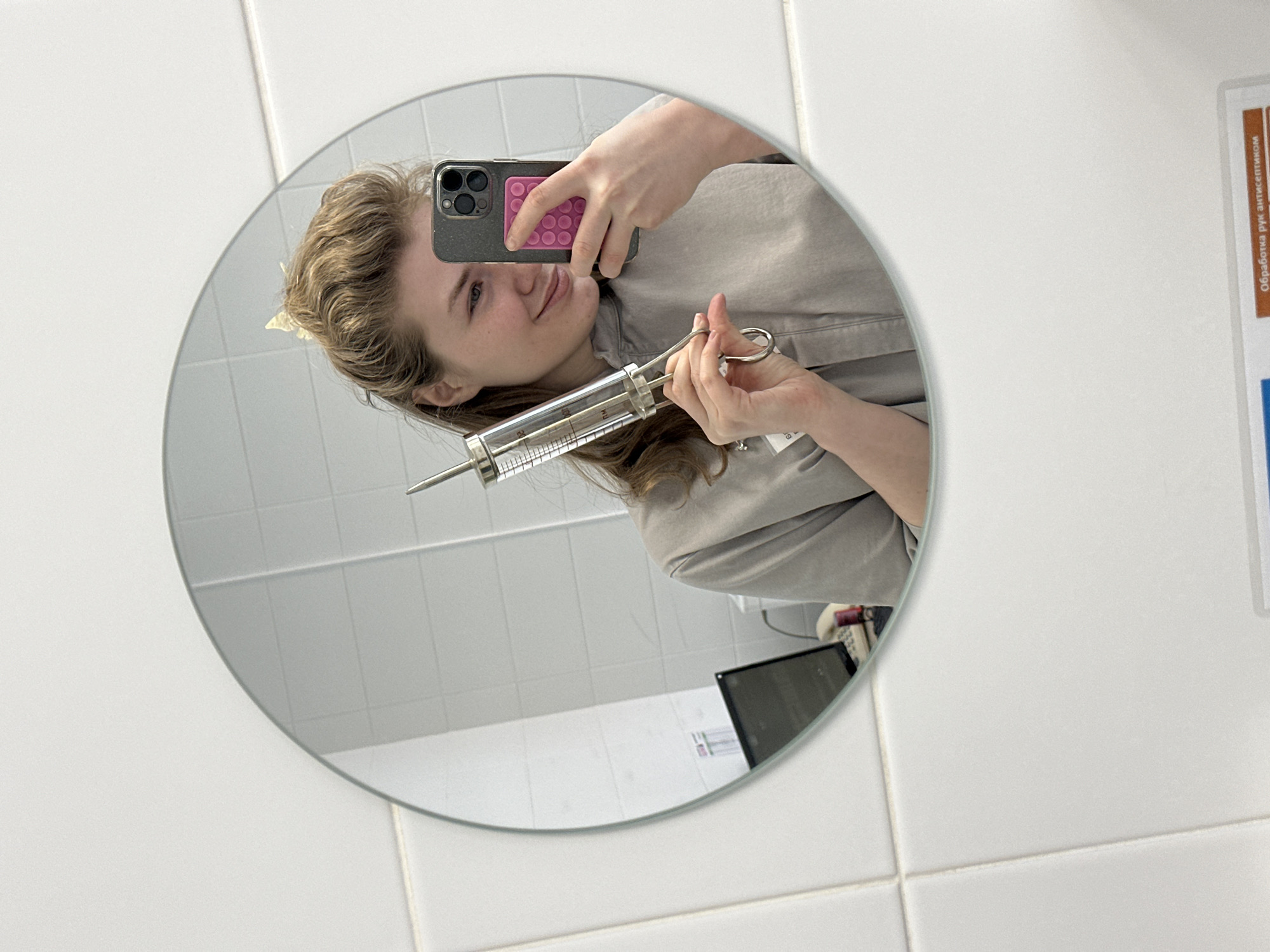 A person with fair skin and wavy brown hair is reflected in a round mirror in what appears to be a tiled bathroom or laboratory setting. The person is holding a large, metal and glass syringe in one hand and a smartphone with a pink case in the other, presumably taking a selfie. The reflection shows part of a document in Russian on the wall to the right of the mirror. A partial view of a dark-colored object (possibly a machine) is visible at the bottom of the mirror's reflection. No information is provided about a specific location in Kazan, Russia, or any specific actions or interactions taking place.