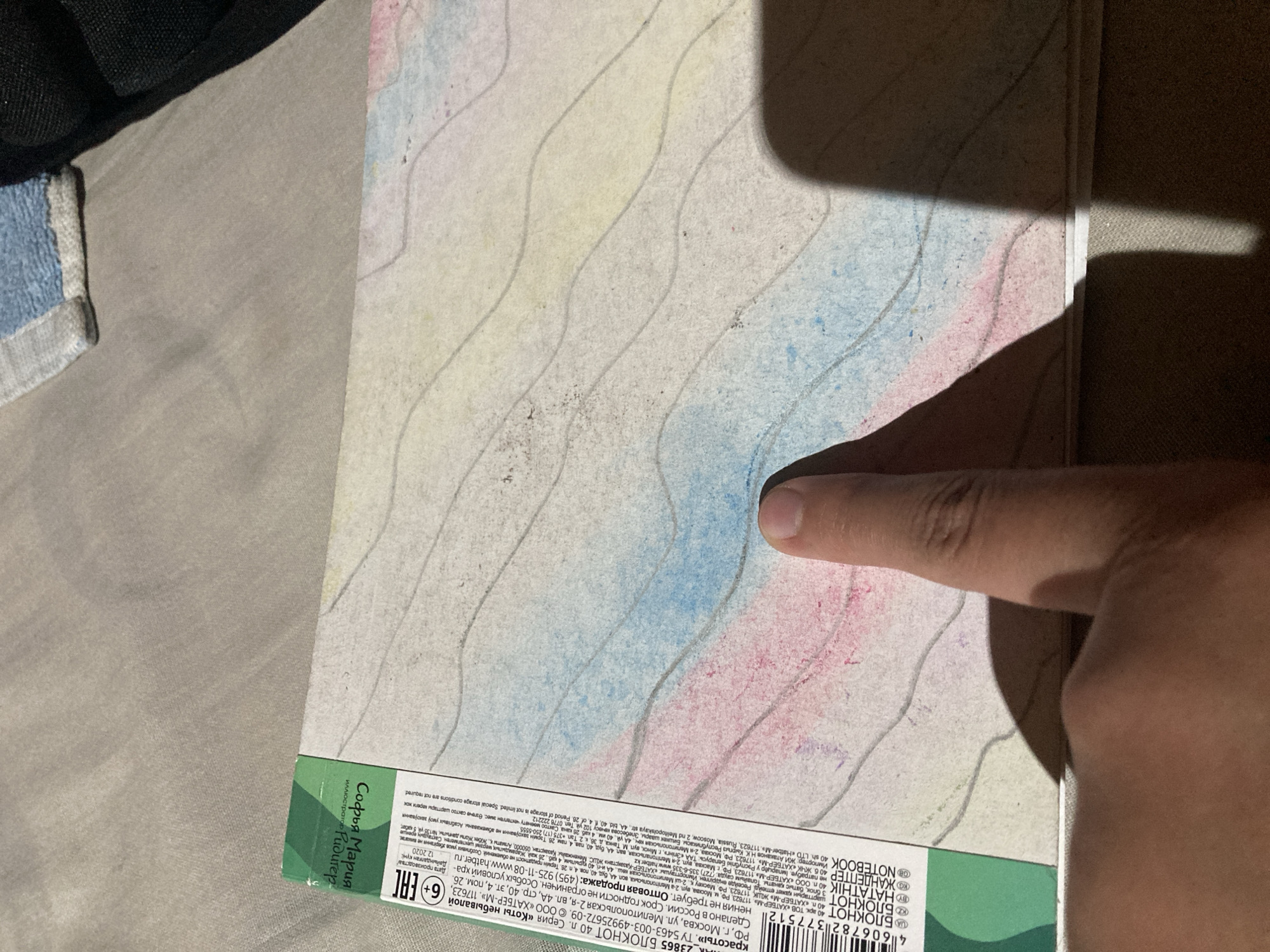 An overhead close-up shot shows a portion of a notebook with a watercolor illustration of wavy lines in blue, pink, and yellow. The notebook's cover is visible in the lower left corner with Russian text and barcodes. A person's thumb is pressing down on the notebook's surface. The notebook is lying on a textured, grey fabric surface, possibly a bed or blanket. The background is blurred, showing more of the grey fabric and a dark object that could be a bag. The text on the notebook indicates it is a "BLOCKNOTE" from the "HATBER" brand and mentions production in Russia. The specific location of Besedy, Russia, is not visually evident in the image, but the text suggests it is the country of origin for the product.