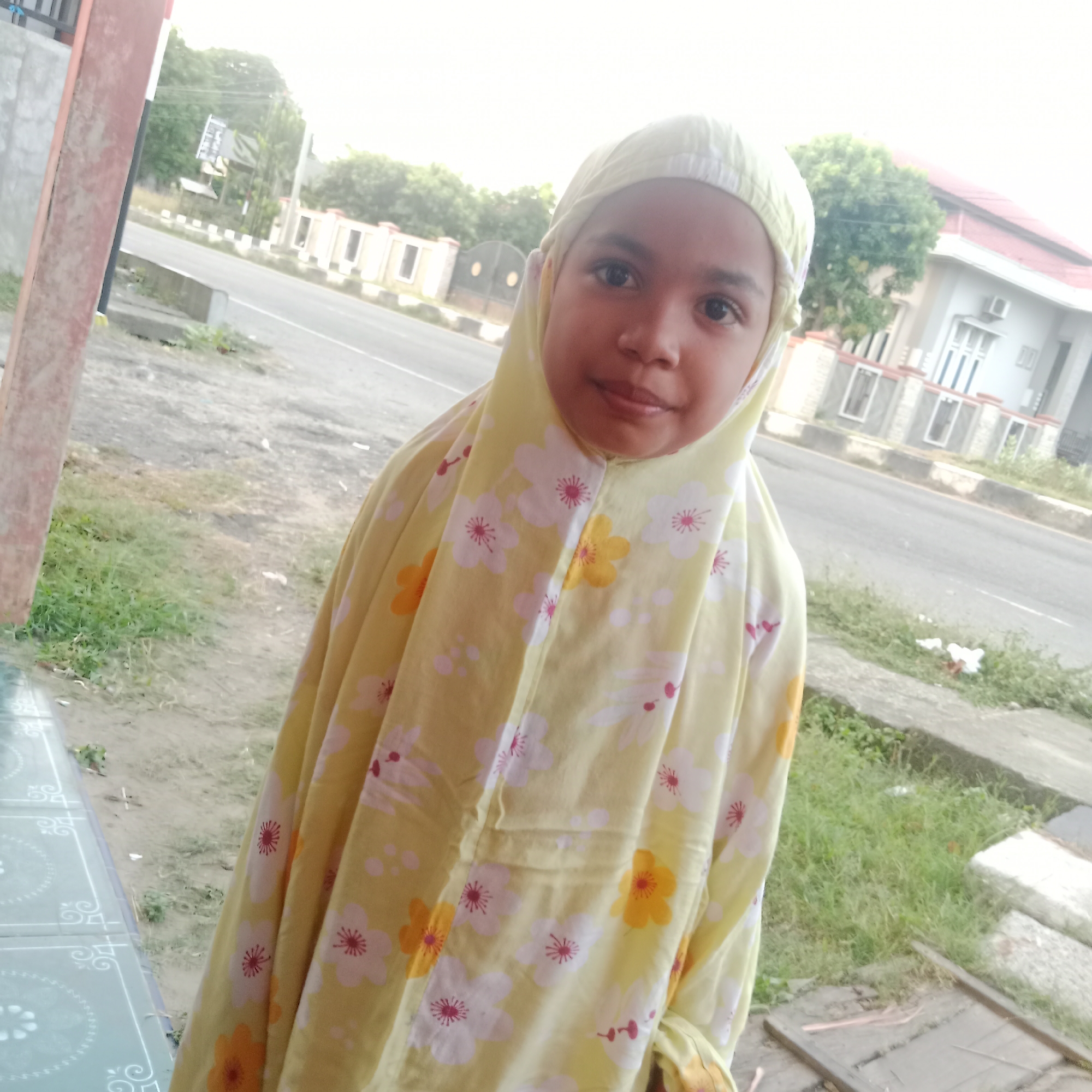A young girl is captured in a daylight portrait, standing outdoors in what appears to be a residential or semi-urban area of Lhokseumawe, Indonesia. She is the central subject, looking directly at the camera with a neutral expression, possibly a faint smile. She wears a light yellow prayer garment (telekung or mukena) that covers her head as a hijab and extends down her body. The garment is adorned with a pattern of white, orange, and red floral motifs.
To her immediate left, a red-brown vertical pillar or structure is partially visible, alongside a light blue-green tiled surface on the ground, possibly part of a veranda or porch. Behind her, a paved road stretches across the mid-ground, flanked by white and black striped barriers or curbs. On the far side of the road, various elements of the urban landscape are visible. These include green trees and other vegetation. To the right, a house with a reddish-brown tiled roof, white walls, and an exterior air conditioning unit is discernible, alongside a fence or wall. Further left across the road, another building or fence structure can be seen, and a distant, indistinct dark sign or billboard with some white markings is barely visible. The ground between the girl and the road features patches of grass, dirt, and some scattered white debris. In the bottom right corner, there are wooden planks and concrete slabs.
The clear, bright lighting indicates it is daytime, likely morning or late afternoon given the soft quality of light. No specific activity beyond posing for the photograph is evident, but her attire suggests a cultural or religious context, possibly related to prayer. No legible text is visible in the image.