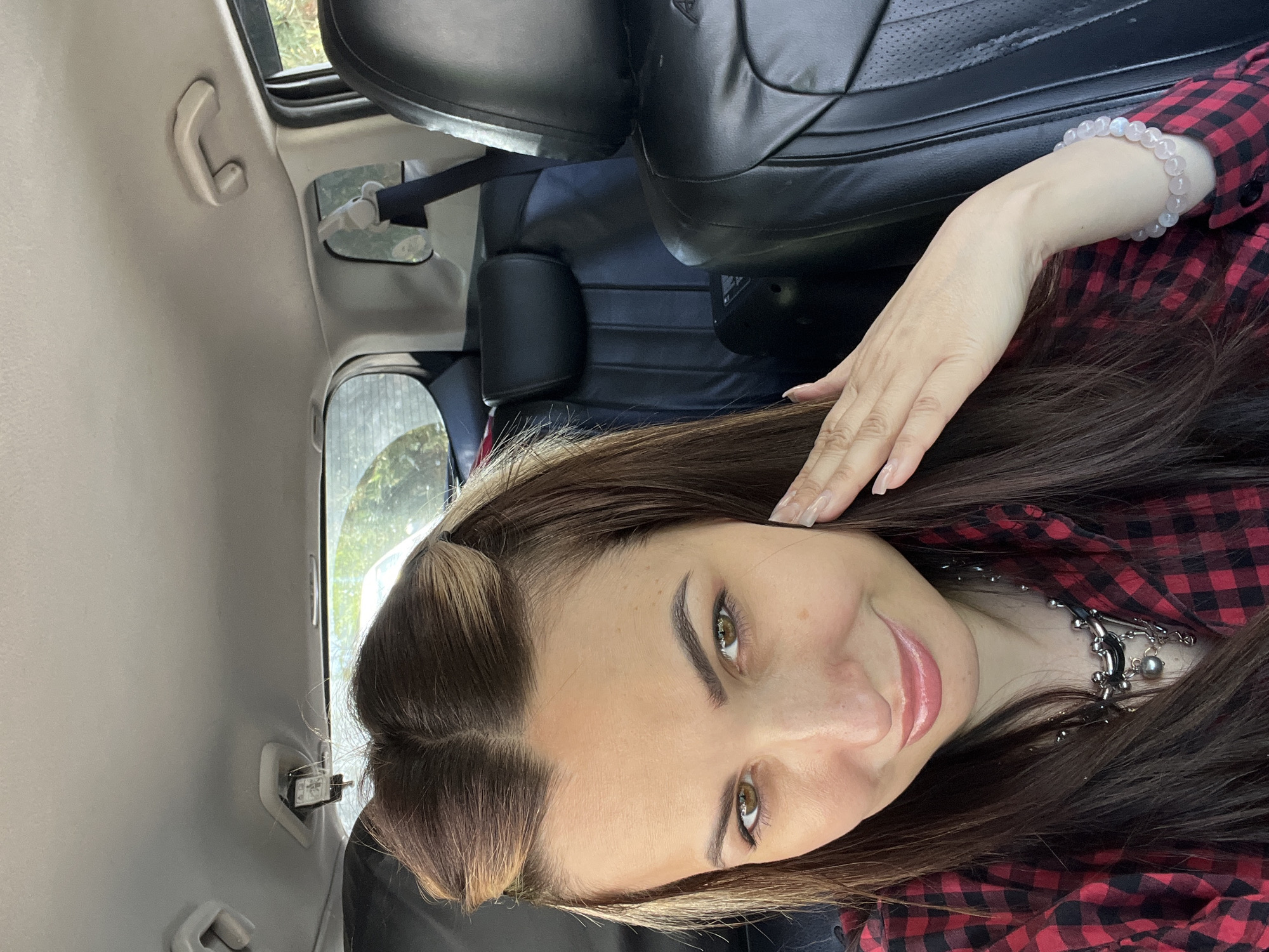 A selfie of a woman with brown hair and green eyes, wearing a red and black plaid shirt and a necklace, is taken inside a car. The car's interior is visible, with black leather seats and grey roof trim. A hand with a bracelet is also visible. The exact location in the city of Unknown, Unknown cannot be determined from the image.