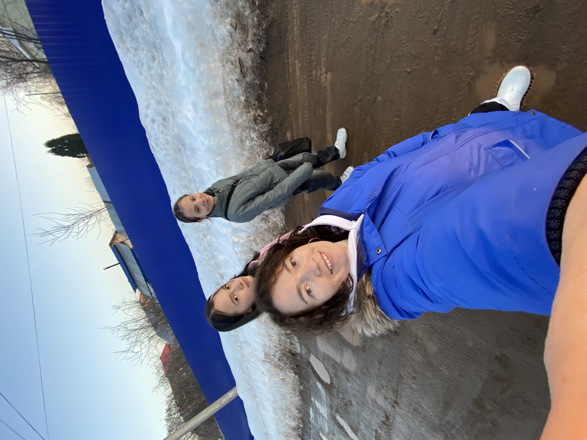 Three females are positioned on a muddy, unpaved road in Ольховка, Russia. In the foreground, a woman with brown curly hair wears a blue winter jacket with a fur-lined hood and a white hoodie, smiling directly into the camera from a low-angle perspective, consistent with taking a selfie. She has a gold hoop earring on her visible ear. Behind her, to the left, a younger female with long dark hair is visible from the chest up, wearing a pink garment and looking towards the camera with a neutral expression. To the right of the second female, a third younger female stands in a grey puffer jacket, dark trousers, and white sneakers, carrying a black shoulder bag, also looking towards the camera with a slight smile. The road surface is brown mud with visible tire tracks. To the immediate right of the subjects, a snowbank with patches of melting snow and ice runs parallel to the road. In the background, a bright blue corrugated metal fence spans horizontally. Beyond the fence, several houses with various roof colors (including red and blue) and a few bare deciduous trees are visible under a clear, light blue sky. A single tall, slender evergreen tree is also present behind the fence on the right side. A utility pole or post is partially visible on the far left. The lighting suggests late afternoon or early morning.