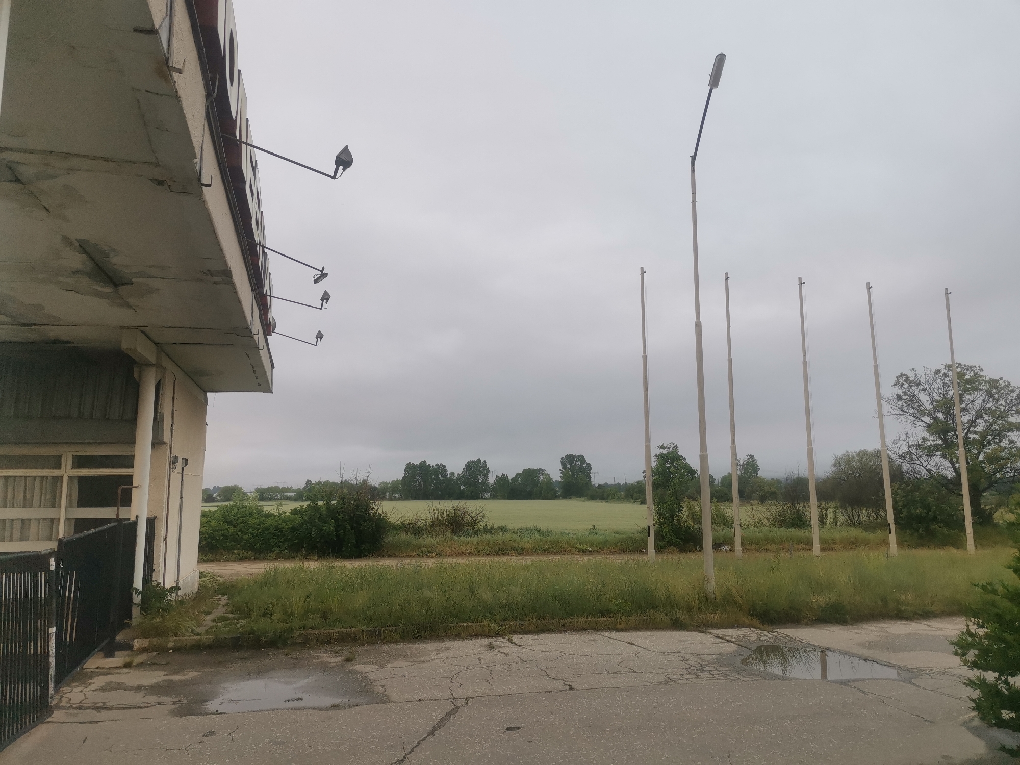 The image captures a quiet, overcast scene, likely in Zhivkovo, Bulgaria, depicting an area that appears to be disused or in a state of neglect. On the left, a weathered concrete building structure is partially visible, featuring a prominent overhang from which several small, angled lights extend, designed to illuminate what was likely once a sign (no legible text remains). The building exhibits staining and wear. A dark metal fence or gate is seen adjacent to the building on the lower left.
The foreground consists of cracked asphalt pavement with several puddles reflecting the grey sky, indicating recent rain. Weeds and tall grass grow abundantly through the cracks and along the edges of the paved area.
To the right, a series of tall, slender poles stands against the horizon. The leftmost of these poles holds a modern street light fixture, while the others are bare or have small, indistinct caps, possibly old flagpoles or supports. These poles are surrounded by overgrown vegetation, including bushes and tall grasses.
Beyond the immediate foreground, a lush green agricultural field stretches into the distance, bordered by a line of trees. The sky is uniformly overcast and grey, diffusing the light and creating a subdued atmosphere, suggesting either a heavily cloudy day or twilight hours.
There are no people or discernible activities taking place, contributing to a sense of stillness and abandonment. The cracked pavement, overgrown vegetation, and weathered structures collectively convey a feeling of decay and natural reclamation in this rural or semi-rural setting.