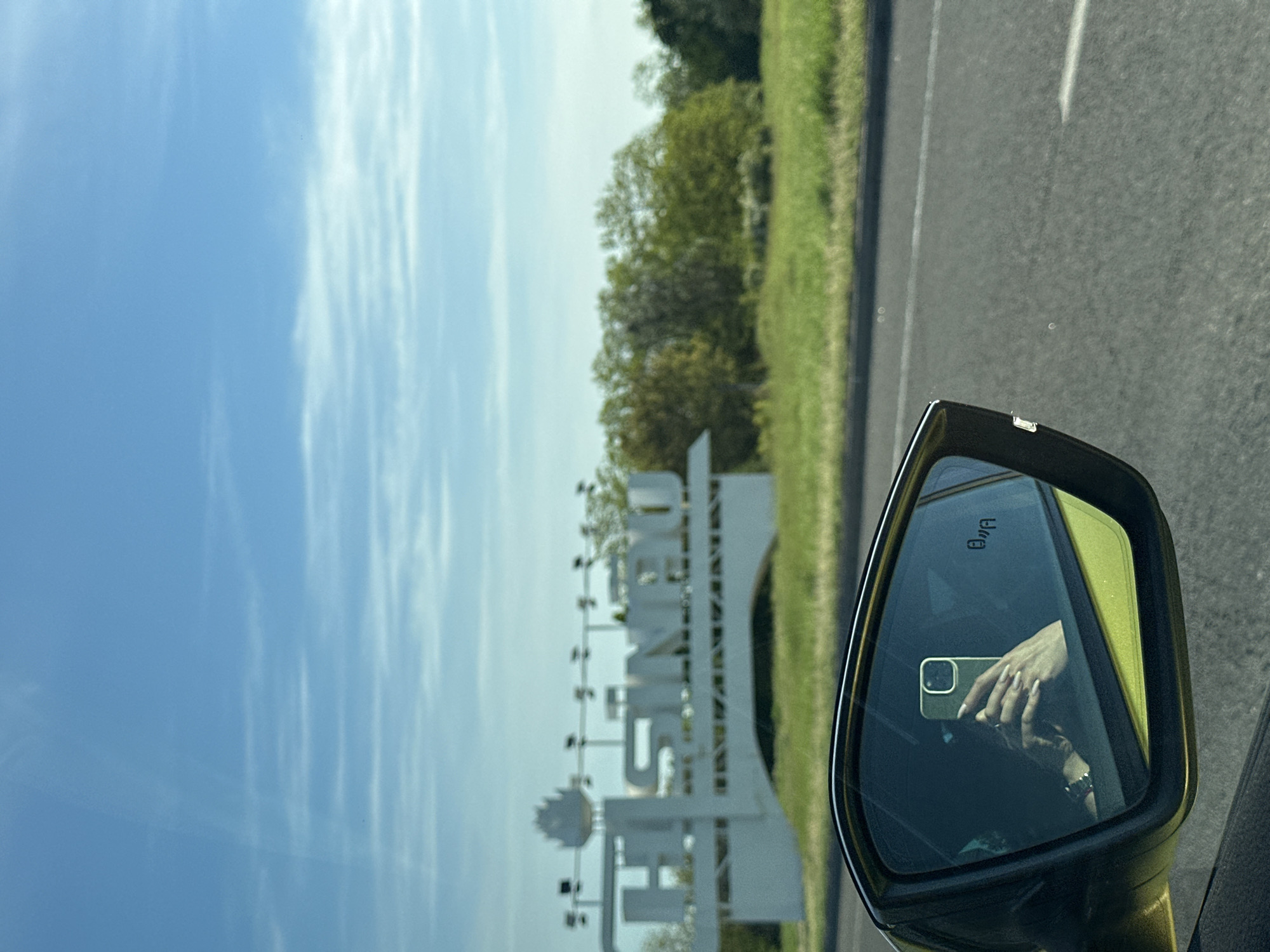The image, taken from inside a vehicle, shows a rearview mirror reflecting a person holding an iPhone. Outside the vehicle, there is a sign that reads "CHISINAU." The setting appears to be a roadway with green vegetation alongside. Based on the sign, the location is near Chișinău. A possible location is the city of Stăuceni, Moldova.