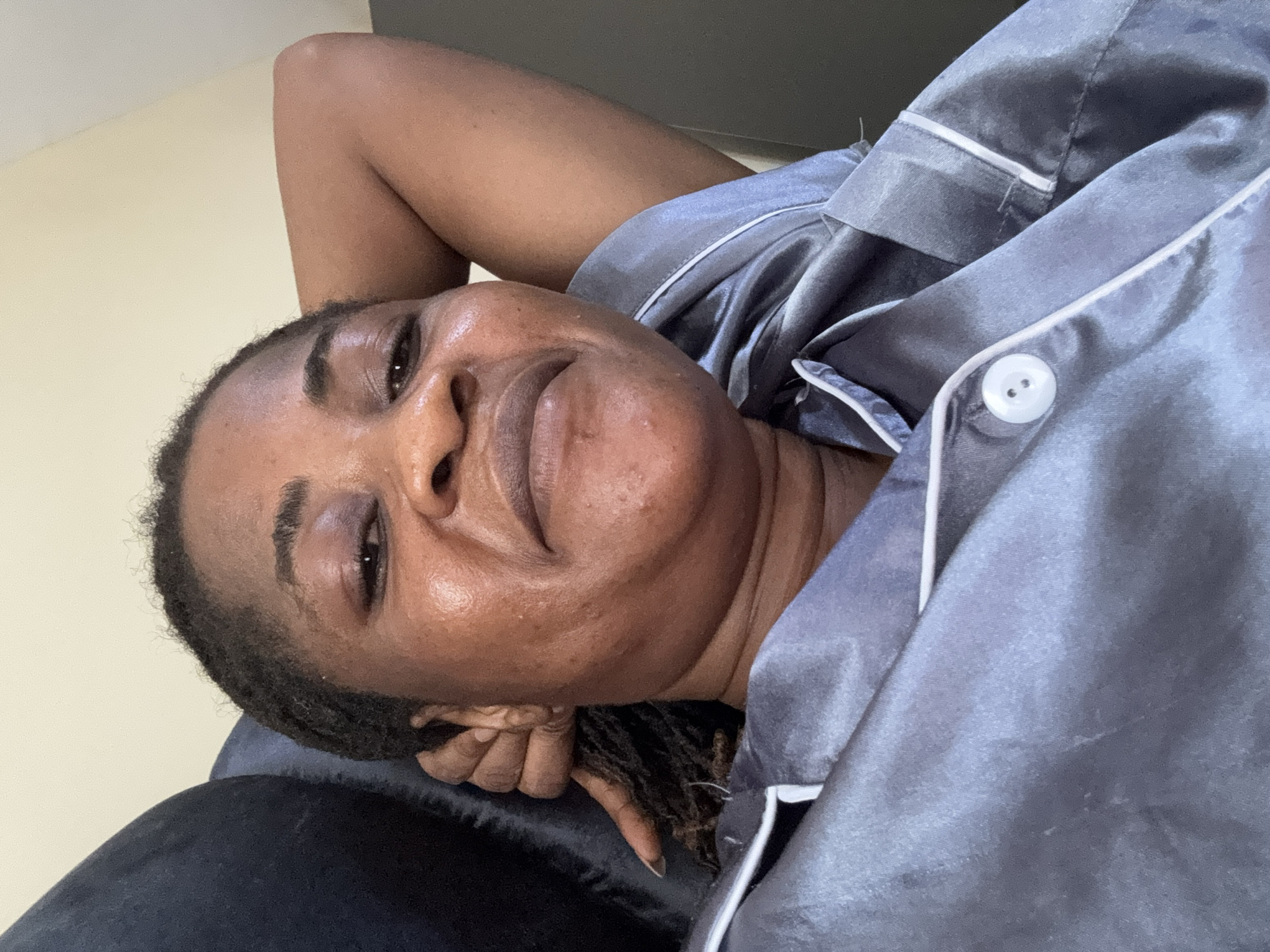 A woman with dark skin, wearing gray pajamas, is shown in a close-up shot. She has her arm behind her head, and she is looking at the camera with a slight smile. The background is a light-colored wall. The image appears to have been taken indoors, possibly in a bedroom, in the city of Ajara, Nigeria.
