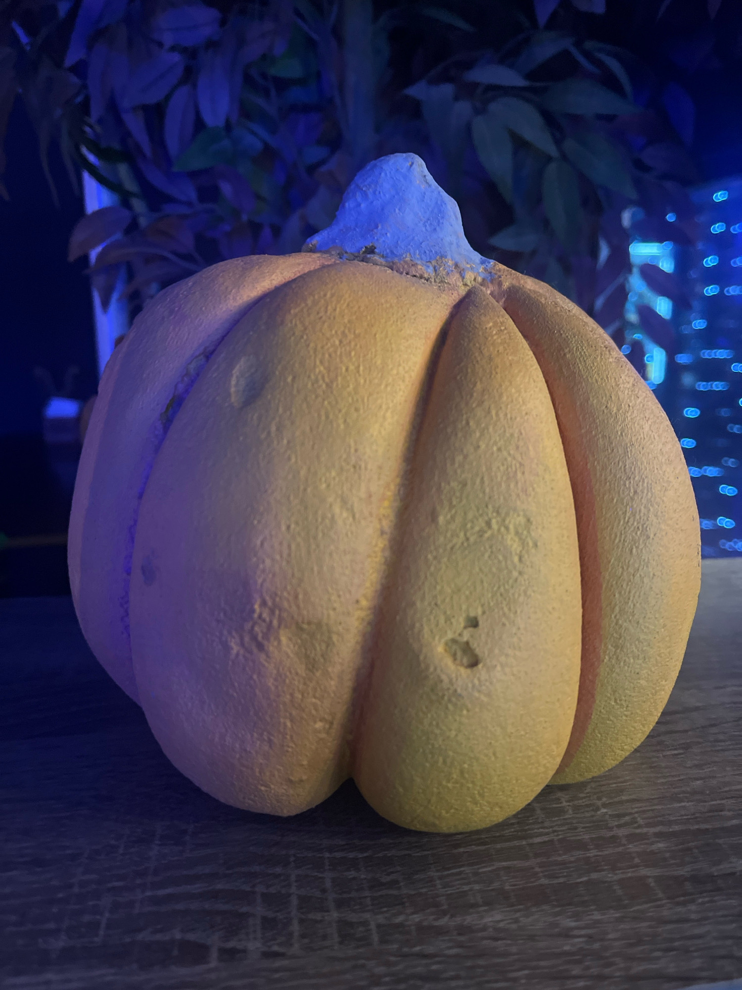 The image shows a close-up of a faux pumpkin, likely for decoration. The pumpkin is orange with distinct ridges and a light blue stem. It is placed on a wooden surface, possibly a table. The background is dark and blurred, with hints of blue and green foliage and some out-of-focus blue lights, suggesting an indoor setting with artificial lighting, possibly a home or a decorated commercial space. There are no people or animals present in the image. The exact location in Shakhty, Russia is not discernible from the visual information provided.