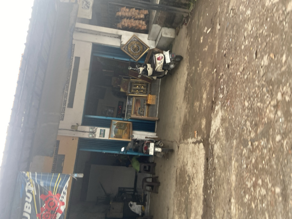 The image shows the exterior of a storefront in Baleendah, Indonesia, located at coordinates -7.021 latitude and 107.656 longitude. Two motorcycles are parked in front of the store, one black and one white. The black motorcycle is partially obscured by a blue door. The white motorcycle is parked in front of a white wall. There is a sign with the word "UBUT" on it. The interior of the storefront is visible, and there are several framed pictures on the walls. Two people are visible in the interior: one is sitting on a chair, and the other is standing. The ground is covered in gravel and dirt, and there are a few empty plastic chairs in front of the store. The building is made of concrete and has a metal roof. There are several hanging objects that appear to be dried fruits. The entrance to the building is open, revealing a large open space. There is a metal door on the right side of the image, and an object is hanging from it. The street is empty, except for the two motorcycles. The sky is overcast and gray. The image is taken from a slightly elevated angle. The image is well lit and in focus.