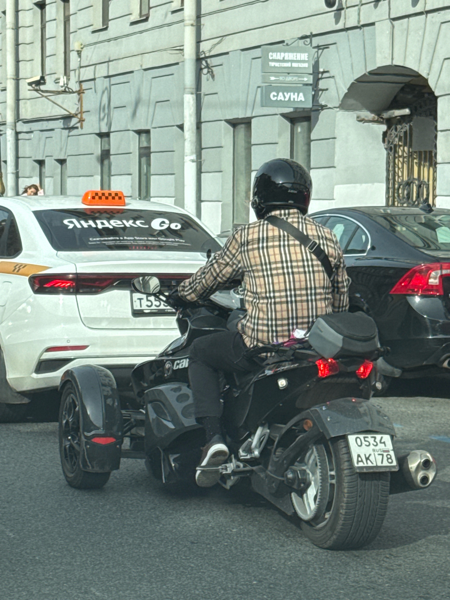 In Saint Petersburg, Russia, a person wearing a checkered shirt and a black helmet rides a black three-wheeled motorcycle. The motorcycle has a license plate that reads "0534 AK 78". The motorcycle is behind a white taxi with an orange checkerboard roof light and the "Yandex Go" logo. In the background, there are buildings with signs that read "CHAPAЖEHME" and "CAVH. The overall scene suggests a street in the city.