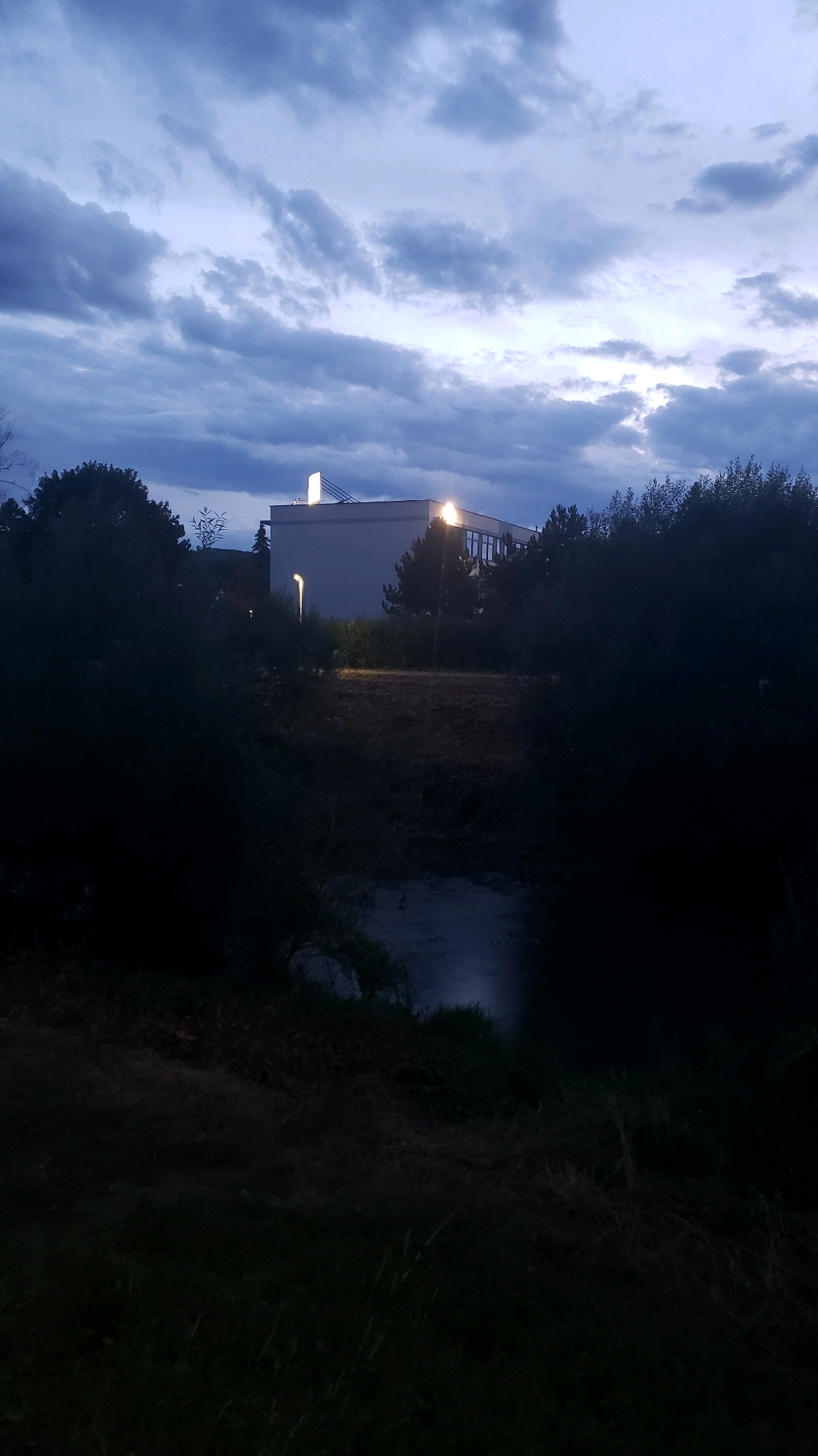 The image depicts a building illuminated by artificial lights against a twilight sky filled with dark, dramatic clouds. In the foreground, a body of water is visible, reflecting the dim light. Dense, dark foliage frames the scene, suggesting a natural or park-like setting bordering a developed area. There are no people visible in the image. The building appears to be a modern structure with windows and a prominent light fixture on its roofline, along with a streetlamp positioned in front of it. The overall atmosphere is one of dusk or early evening. This scene is located in Prešov, Slovakia.