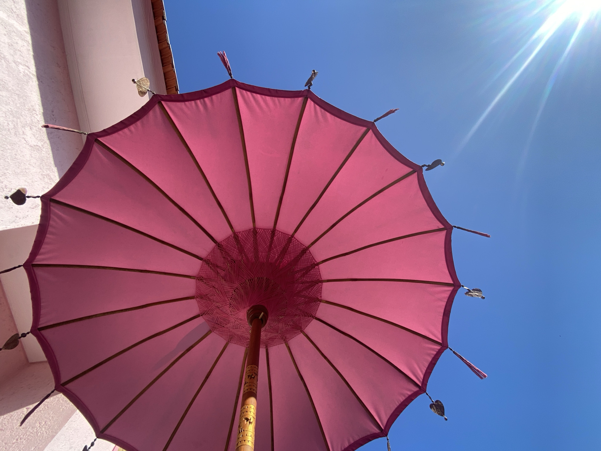 An overhead, low-angle shot shows a bright pink umbrella against a clear blue sky. The umbrella is a decorative parasol, with wooden ribs supporting the fabric. Several decorative tassels and metal heart-shaped charms hang from the edge of the umbrella. The umbrella is attached to a wooden pole, which is carved with decorative patterns. In the upper left corner, a portion of a ceiling or overhang is visible, painted a light pink hue. Sunlight flares brightly in the lower right corner of the image, suggesting it is a sunny day. The scene is static, with no visible movement or people.