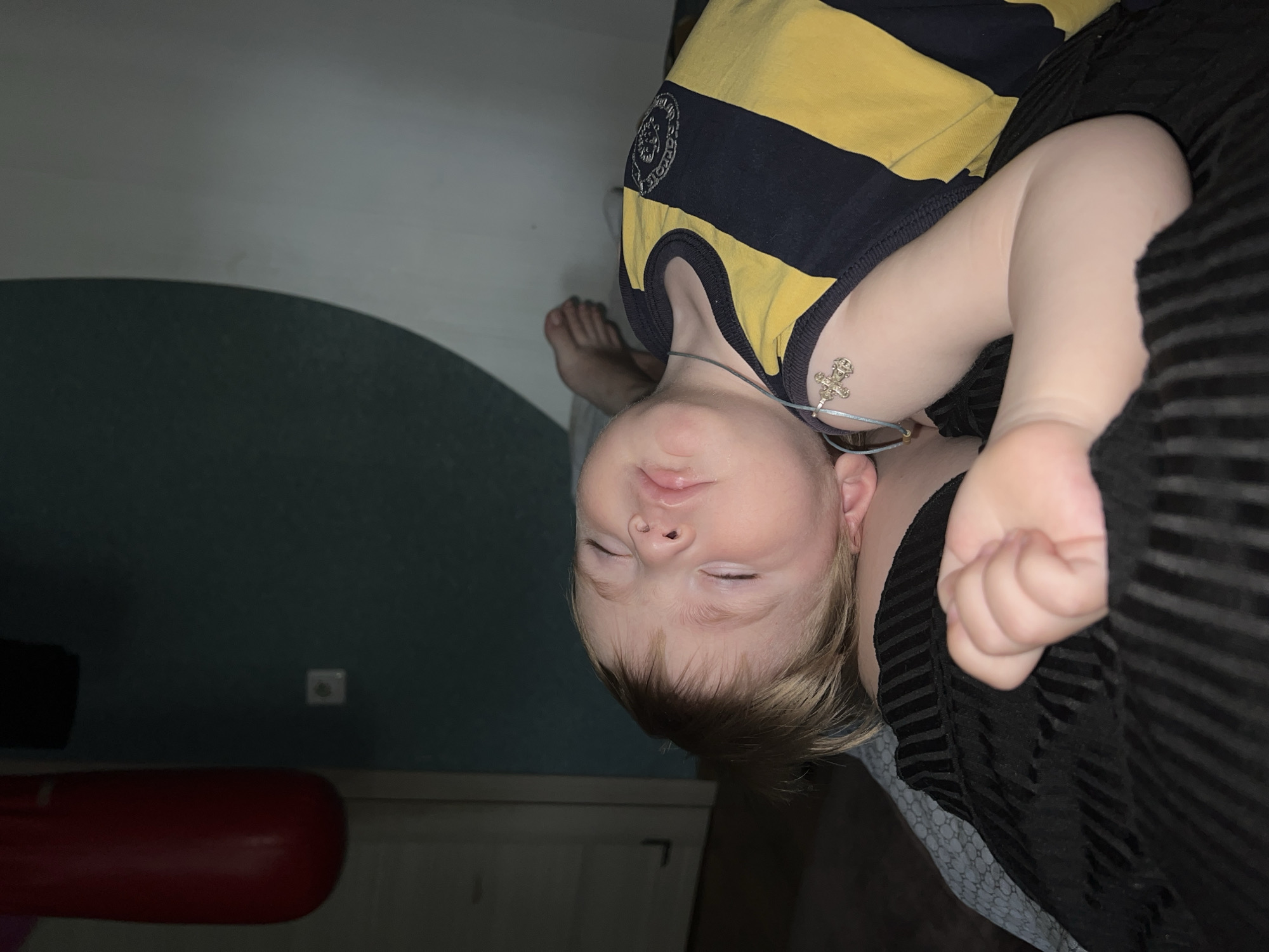 A young child is sleeping indoors. The child is wearing a yellow and blue striped shirt and a necklace with a cross pendant. The child's right hand is clenched. A striped black garment is draped over part of the child. In the background is a teal-colored wall with an electrical outlet, a red cylindrical object, and a white door or drawer. There is no indication of this image being in Noviy, Russia.
