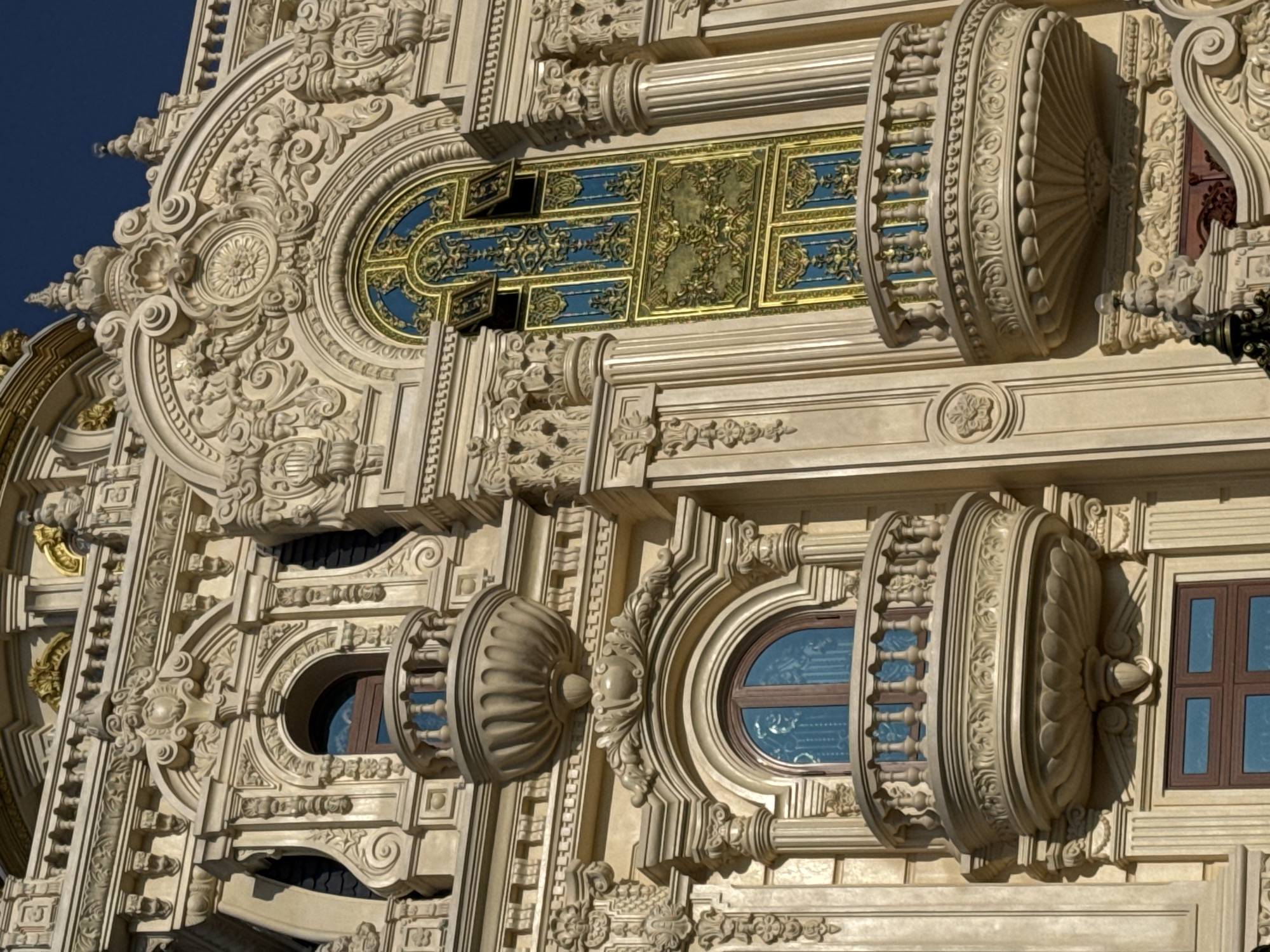 The image displays an exterior view of a building with ornate, baroque-inspired architecture. The facade is richly decorated with sculpted elements, including columns, cornices, and intricate carvings. Several balconies with detailed railings and sculpted supports are visible, along with arched windows framed by elaborate ornamentation. One prominent window features a multi-paned design with blue and gold accents. The building's material appears to be a light-colored stone or stucco, with some metallic gold detailing on the upper levels. The sky is clear and blue. This building is the Crazy House, also known as Hằng Nga Guesthouse, located in Da Lat, Vietnam. There are no people or animals visible in the frame.