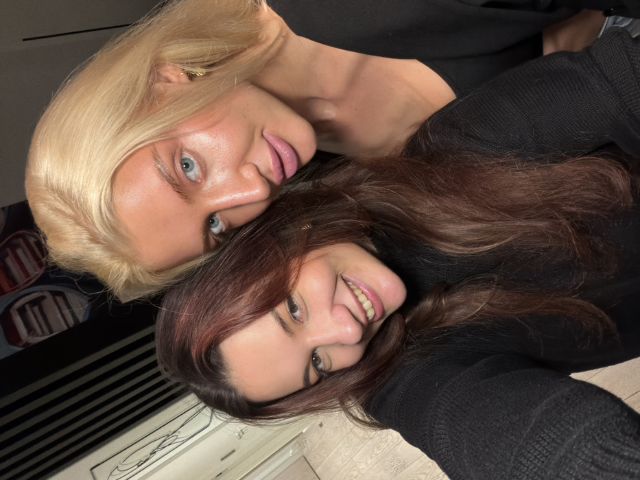 Two women are captured in a close-up, angled selfie, appearing to be lying down or with the camera tilted. Both are looking directly at the viewer with warm expressions.
The woman in the upper portion of the frame has striking blonde hair, styled to fall around her head. She has light blue eyes, a clear complexion with a visible tan, and full lips with a pinkish tint. She is wearing a dark-colored top, possibly black. A small, subtle gold earring is visible in her right ear. Her face is positioned slightly above and to the left of the other woman's head.
The second woman, positioned in the lower and right portion of the frame, has long, wavy brown hair with subtle reddish highlights. She has dark eyes and a wide, genuine smile, revealing her teeth. She is also dressed in a dark, possibly black, long-sleeved top with a visible knit texture on the sleeve. Her head is nestled close to the blonde woman's, suggesting a close relationship.
The background is softly blurred but provides hints of an indoor setting. To the left of the blonde woman's head, there's a dark, patterned object, possibly a cushion or piece of furniture. Below the brown-haired woman, a light-colored floor with some pattern, possibly tiles or wood, is visible. Further in the background, there are dark horizontal lines and a white, perhaps wooden, structure that resembles a cabinet or drawer. The lighting appears soft and warm, highlighting the women's faces. The overall mood of the image is joyful and intimate.