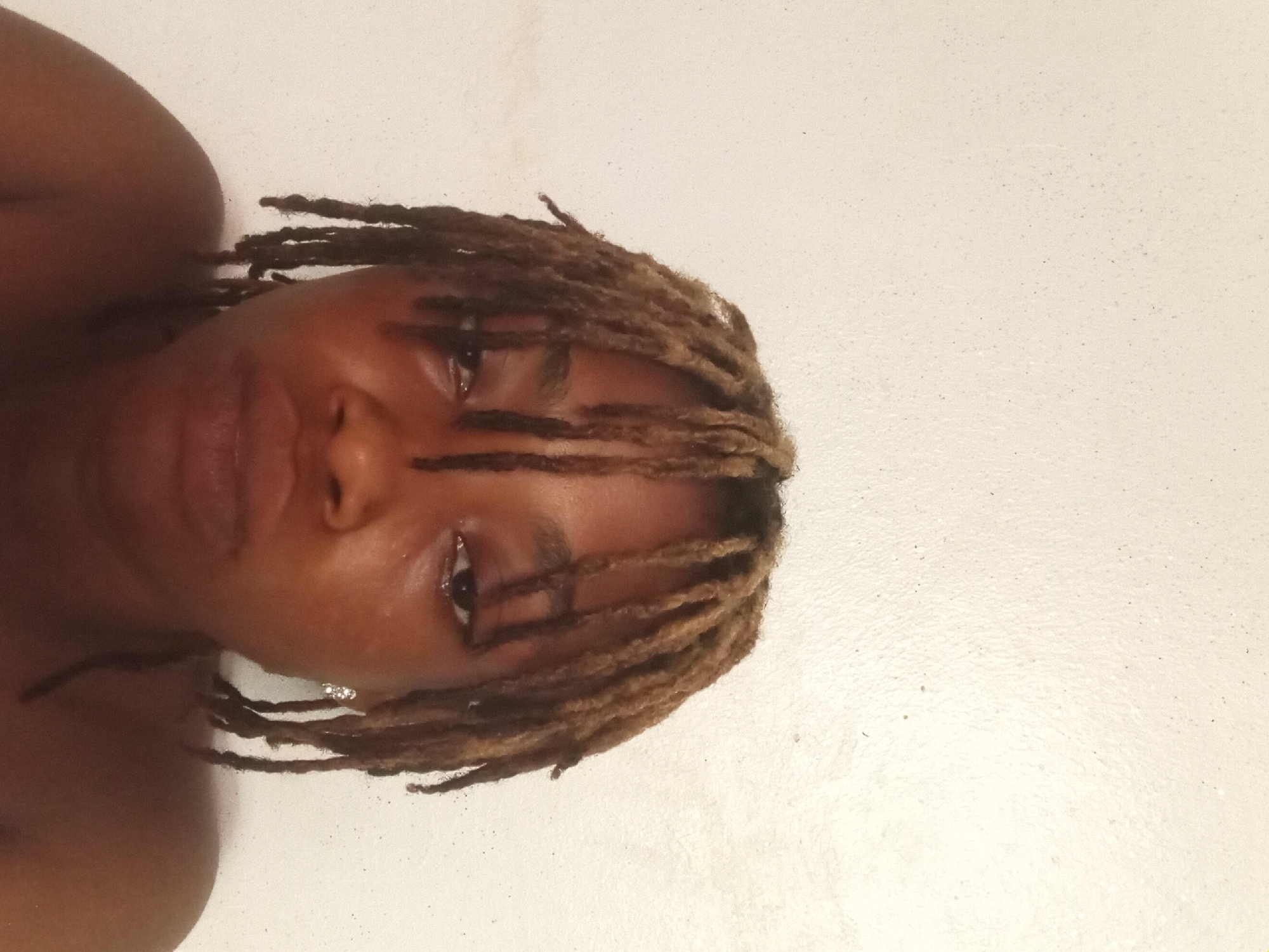 A close-up, portrait-style photograph shows a Black person with brown locs looking directly at the camera. The subject's face is centered in the frame, with the top of the head near the upper edge. The background is a white surface, possibly a wall or other plain backdrop. The person is wearing an earring in their left ear. The image's orientation is slightly rotated, with the head tilted to the left. The location is listed as Unknown, Unknown.