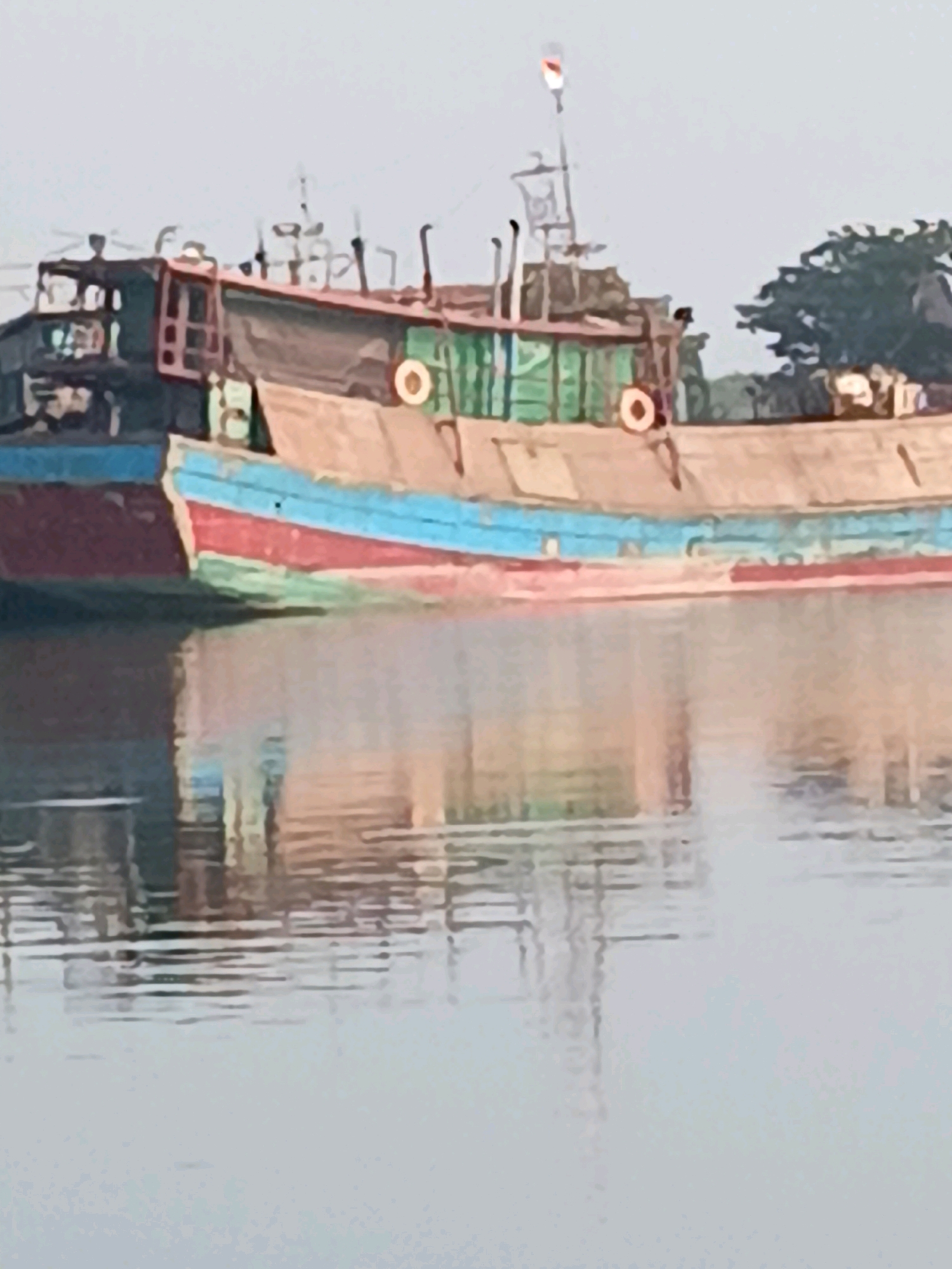 The image depicts a moored cargo ship with a tan and green hull, and a red and blue stripe along the waterline. The ship appears aged and weathered. In the background, a few trees are visible, and the sky is overcast and pale. The ship is reflected in the calm water it is floating on. The scene suggests a port or harbor environment. The ship is located in Cirebon, Indonesia.