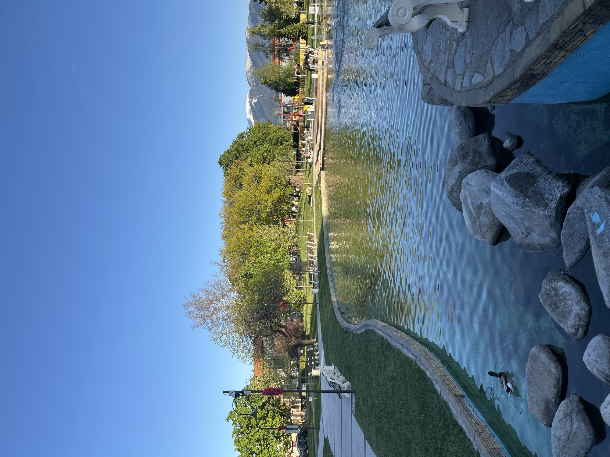 The image depicts a park scene with a pond in Unknown, Unknown. The pond occupies the right side of the frame, featuring rippling water and several large rocks along its edge. A duck swims near the rocks. A grassy bank separates the pond from a paved walkway lined with benches and lampposts. Beyond the walkway, there are trees with varying foliage, some in full leaf and others partially bare. In the background, a playground is visible, with distant mountains completing the landscape. The sky is clear and blue.