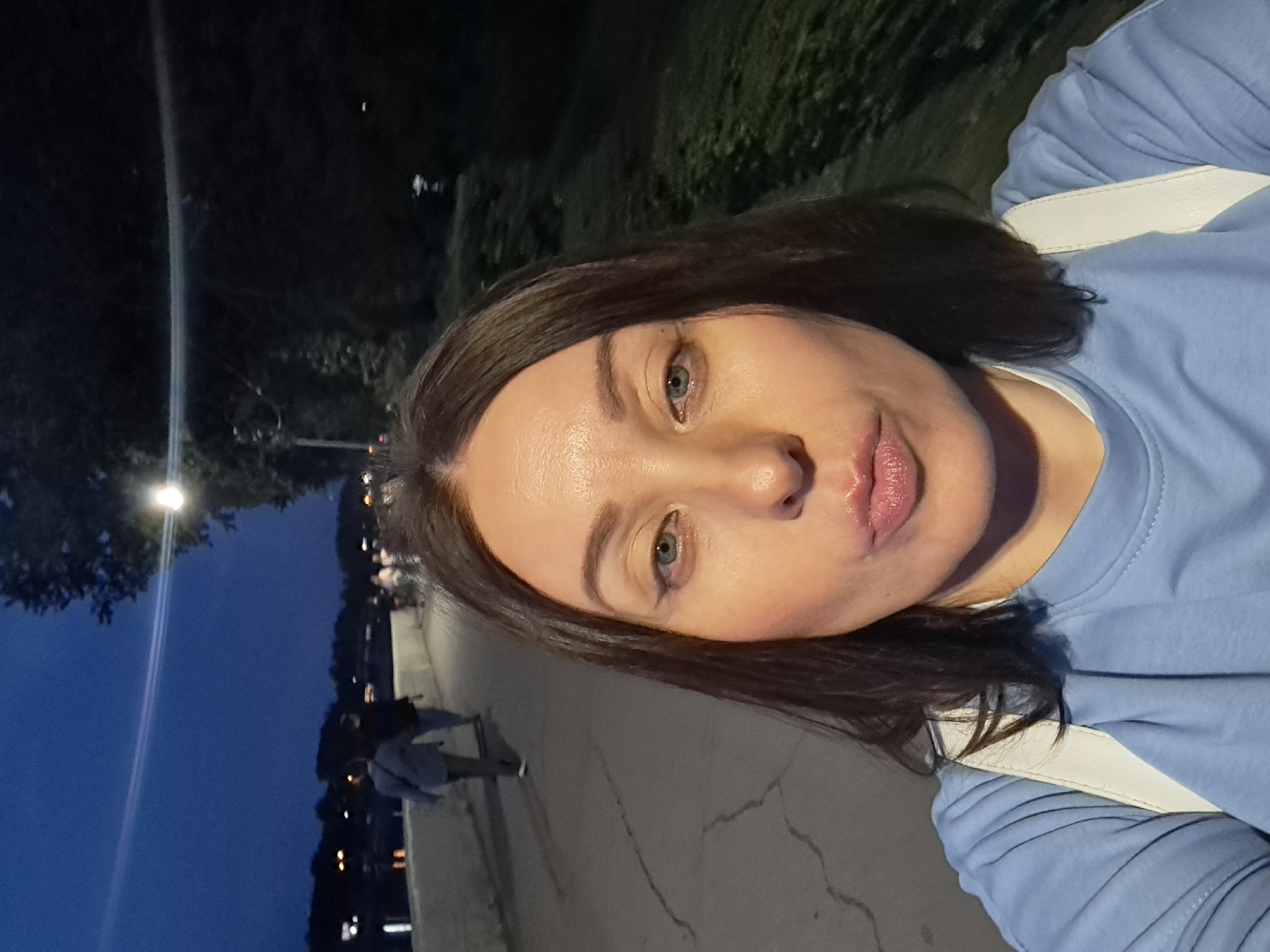 In Irkutsk, Russia, at dusk, a woman is taking a selfie in a public park. She is wearing a light blue t-shirt and a white backpack. The background shows a paved walkway with a few people strolling in the distance. A lamppost illuminates the scene, casting shadows on the path. Trees and grass line the side of the walkway, and lights from across a body of water are visible in the far distance. The woman is looking at the camera with her lips pursed.