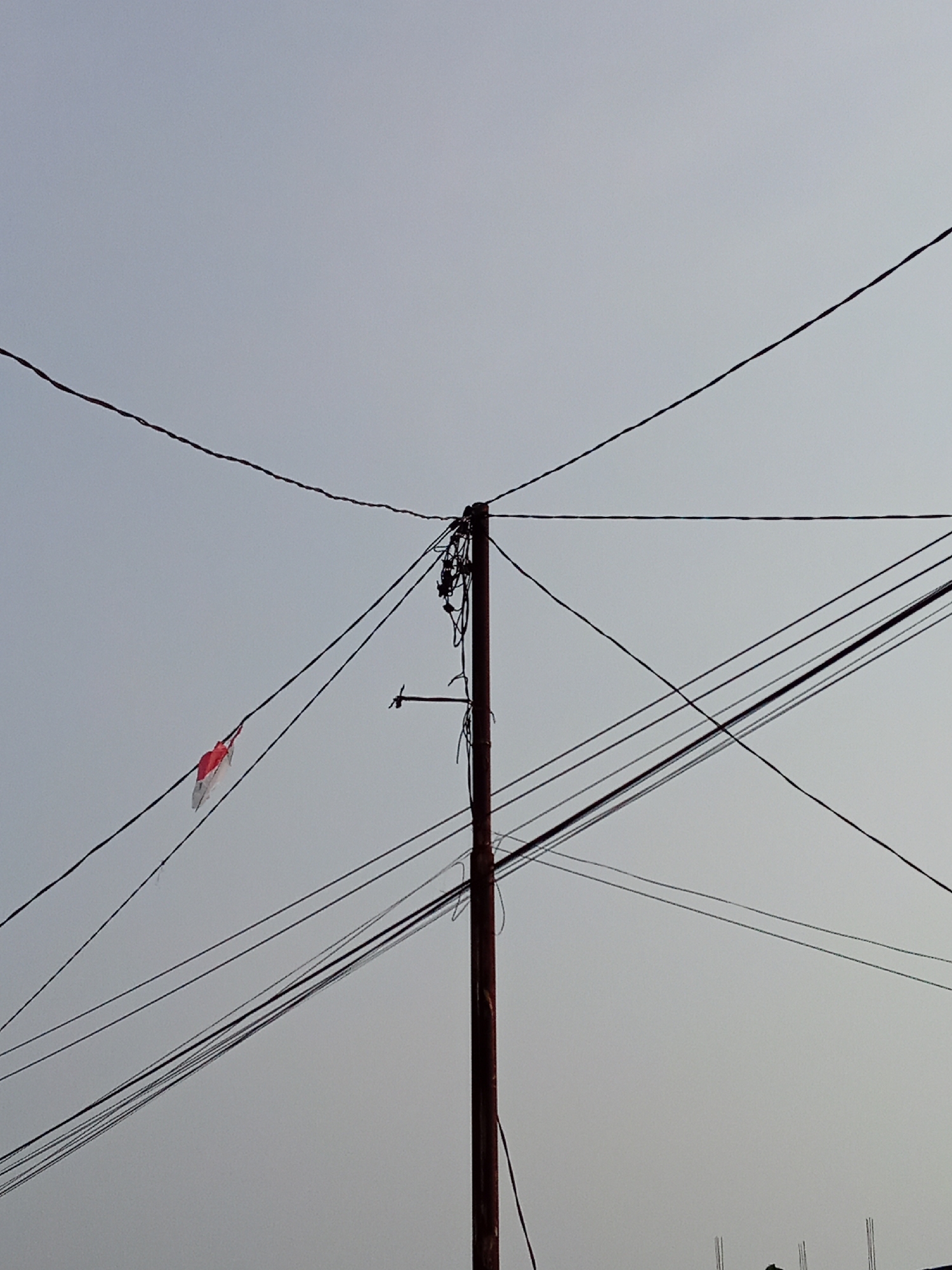 In the city of Tanah Mas, Indonesia, a weathered wooden utility pole stands against a pale, overcast sky, supporting a complex network of dark electrical wires. These wires crisscross the frame at various angles, some taut and others slack, creating a tangled web. A small, tattered flag, predominantly red and white, is caught and hanging from one of the wires on the left side of the pole, its fabric appearing torn and faded. The overall scene is one of utility infrastructure in a modest urban environment under a diffused light.