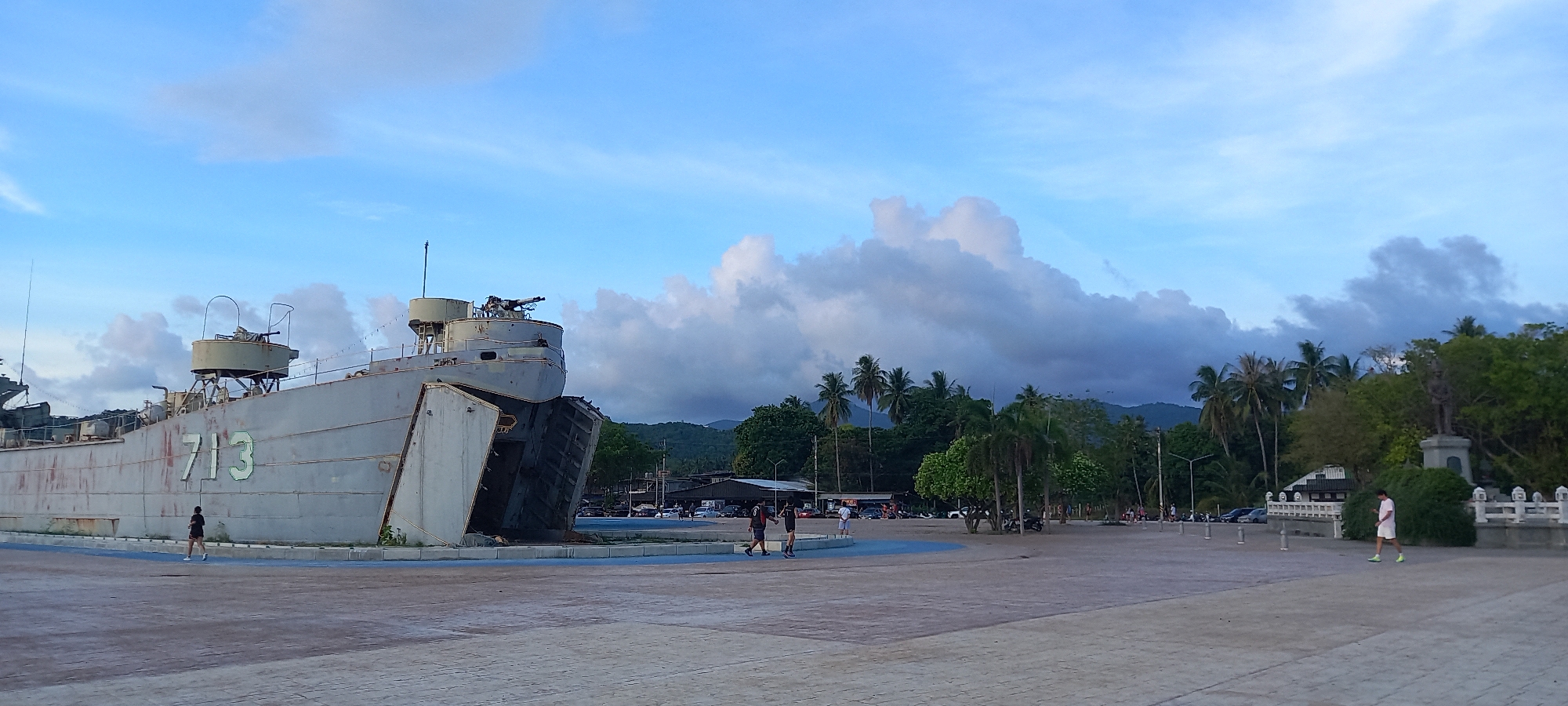 The image depicts a decommissioned World War II Landing Ship Tank (LST) 713 situated in Thong Sala, Thailand. The ship is painted gray with the hull number "713" prominently displayed. It appears to be a museum or memorial exhibit, as it's placed on a paved area with a blue base surrounding it. Several people are visible in the scene. One individual is jogging near the ship. Two people are walking together in the middle ground, and another person is walking along a sidewalk or edge of the paved area in the foreground. The background includes a combination of palm trees, other trees, buildings, and possibly parked vehicles. The sky is partly cloudy, indicating either early morning or late afternoon lighting conditions. Mountains can be seen in the distance behind the trees.