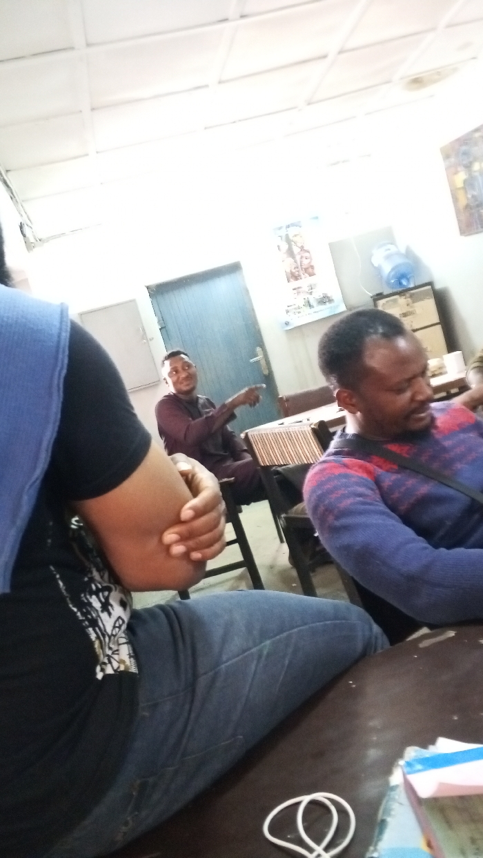 The image depicts an indoor gathering in the city of Dakata, Nigeria. Three individuals with dark skin are visible, seated at dark wooden tables. In the immediate foreground, partially obscured, is a person wearing a black t-shirt under a blue sleeveless top and blue jeans, with their right arm bent. In the mid-ground, a man in a dark maroon long-sleeved tunic is seated, smiling and pointing his right index finger to the right. To his right, another man in a blue, red, and white striped long-sleeved sweater is seated, looking downwards and to his right.
The environment features light-colored walls and a white grid-patterned ceiling with fluorescent lighting. A light blue door with a metallic handle is visible on the left side of the background. A poster featuring human figures is affixed to the wall behind the man in maroon. On the right, a blue water dispenser bottle rests on a cabinet or shelf, next to a partially visible framed item on the wall. A white coiled cable and a stack of papers with a blue cover are present on the foreground table. The seating arrangement suggests a meeting, classroom, or communal space.