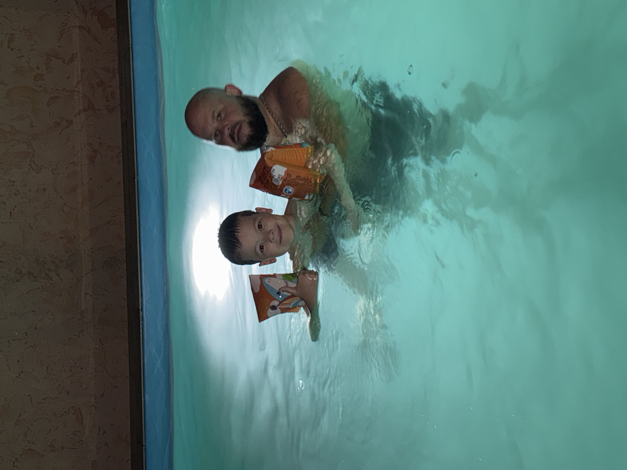 A man and a boy are in a swimming pool. Both are wearing swim floaties. The man has a beard and is behind the boy. The pool has light green water. The side of the pool is bordered by blue tiles, next to which is a textured brown wall. The photo was taken in Krasnaya Polyana, Russia.