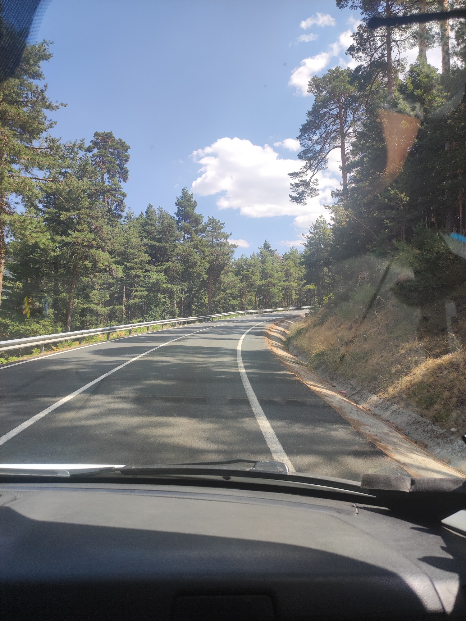 The image depicts a view from inside a vehicle, looking out through the windshield onto a paved road winding through a forest. The road has distinct white lane markings. On the left side of the road, a silver metal guardrail runs parallel to the asphalt. The road is bordered by a dense forest of tall pine trees. The sky above is blue with scattered white clouds. The foreground shows the dashboard of the vehicle. The scene suggests a drive through a natural, wooded area. The specific location of Real Sitio de San Ildefonso, Spain, is not visually evident within the image itself, but it is provided as context.