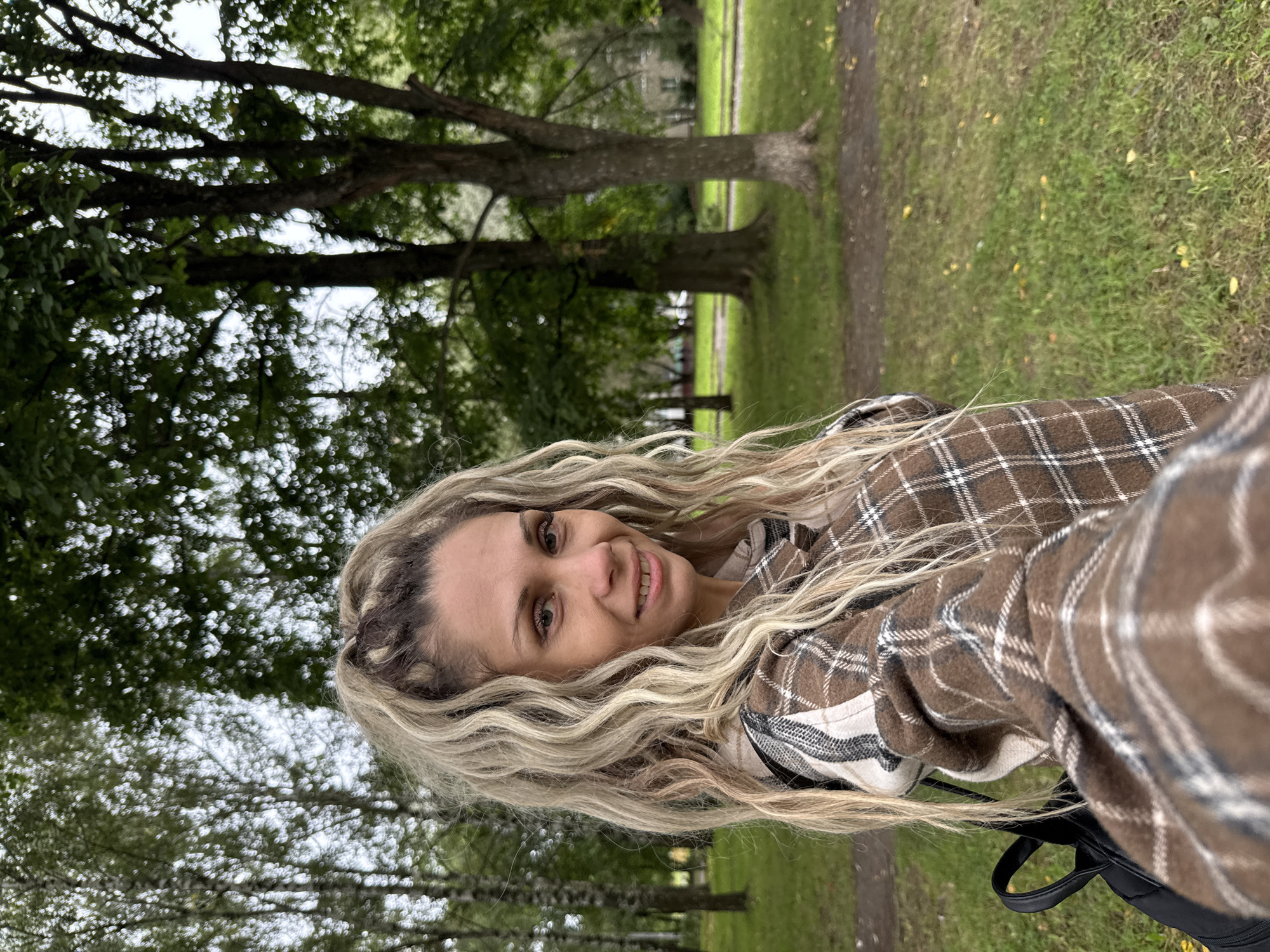 In Rusanovo, Russia, a woman with long, curly blonde hair and wearing a brown plaid jacket poses for a selfie in a park. She is smiling at the camera. The background shows trees with green foliage and a grassy area, suggesting an outdoor setting. A black backpack is visible over her shoulder.