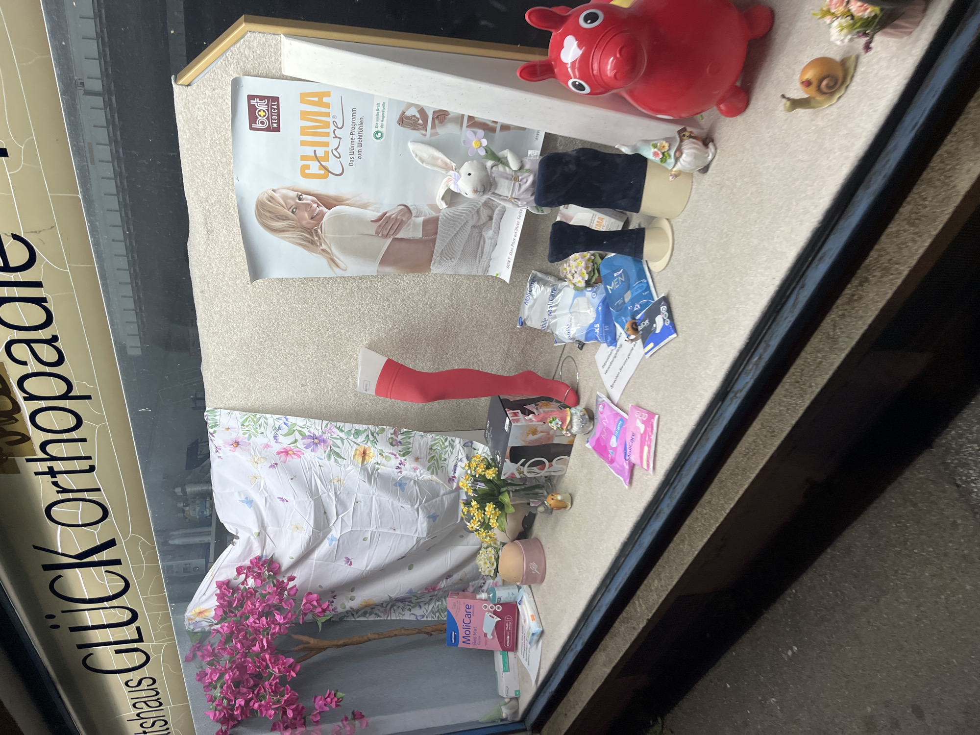 The image shows a shop window display for "GLÜCK Orthopädie" in an unidentified city. A magazine or promotional material titled "CLIMA Care" by "bort MEDICAL" is prominently displayed, featuring a woman and text in German. Other items in the window include a red toy, a stuffed bunny, and medical products, notably from the "MoliCare" line, like "Bed Mat" and XS-sized products. A red stocking on a leg form, a floral-patterned cloth, and artificial flowers add to the display. The scene captures a mix of medical, promotional, and decorative elements within the shop window.