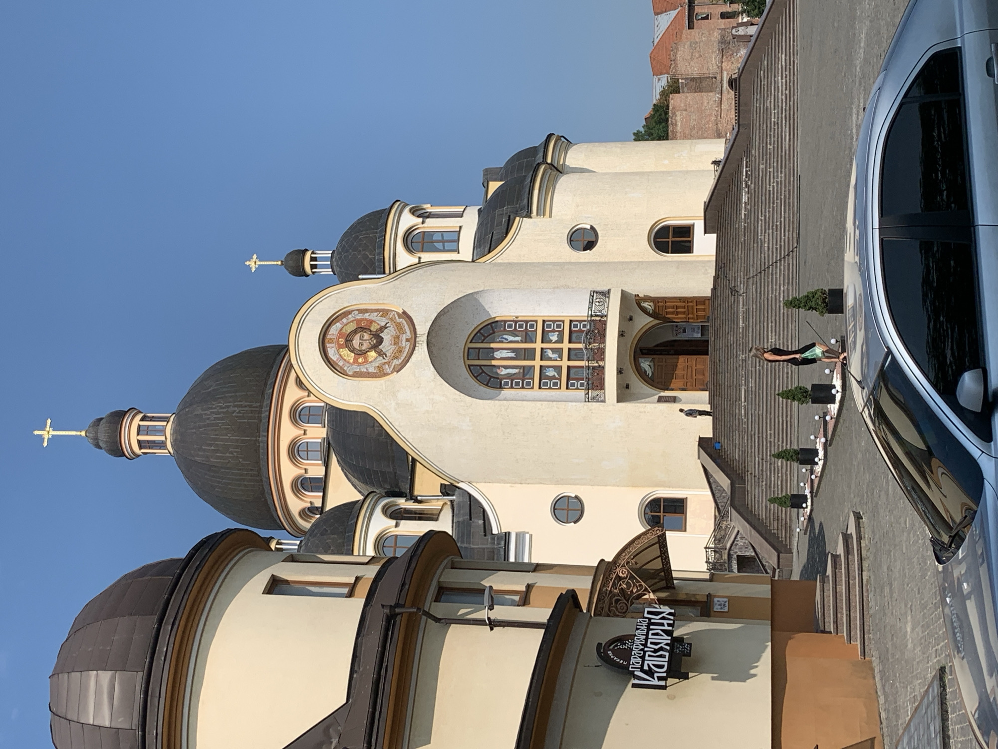 The image depicts the exterior of the Church of the Most Sacred Heart of Jesus in Kolomyia, Ukraine, on a clear, sunny day. The church features a large central dome topped with a golden cross, and multiple smaller domes, all dark in color. The facade is cream-colored with arched windows and decorative mosaic work. A prominent mosaic of Jesus' face is located above the main entrance.
To the left of the church, a building with a brown and cream exterior is visible, featuring a sign with Cyrillic lettering that reads "ПАРАФІЯЛЬНА КАВ'ЯРНЯ" (Parish Cafe). A metal awning with ornate detailing extends over the cafe entrance.
A wide set of stairs leads up to the church entrance, flanked by decorative planters with small evergreen trees. Several people are present: a woman in a black dress walking down the stairs on the right side of the frame, and a person standing near the top of the stairs on the right. A silver car is parked in the foreground, partially obscuring the view of the paved area in front of the church. In the background, the warm brick tones of other buildings are visible, suggesting a developed urban environment. The sky is a clear, bright blue.
