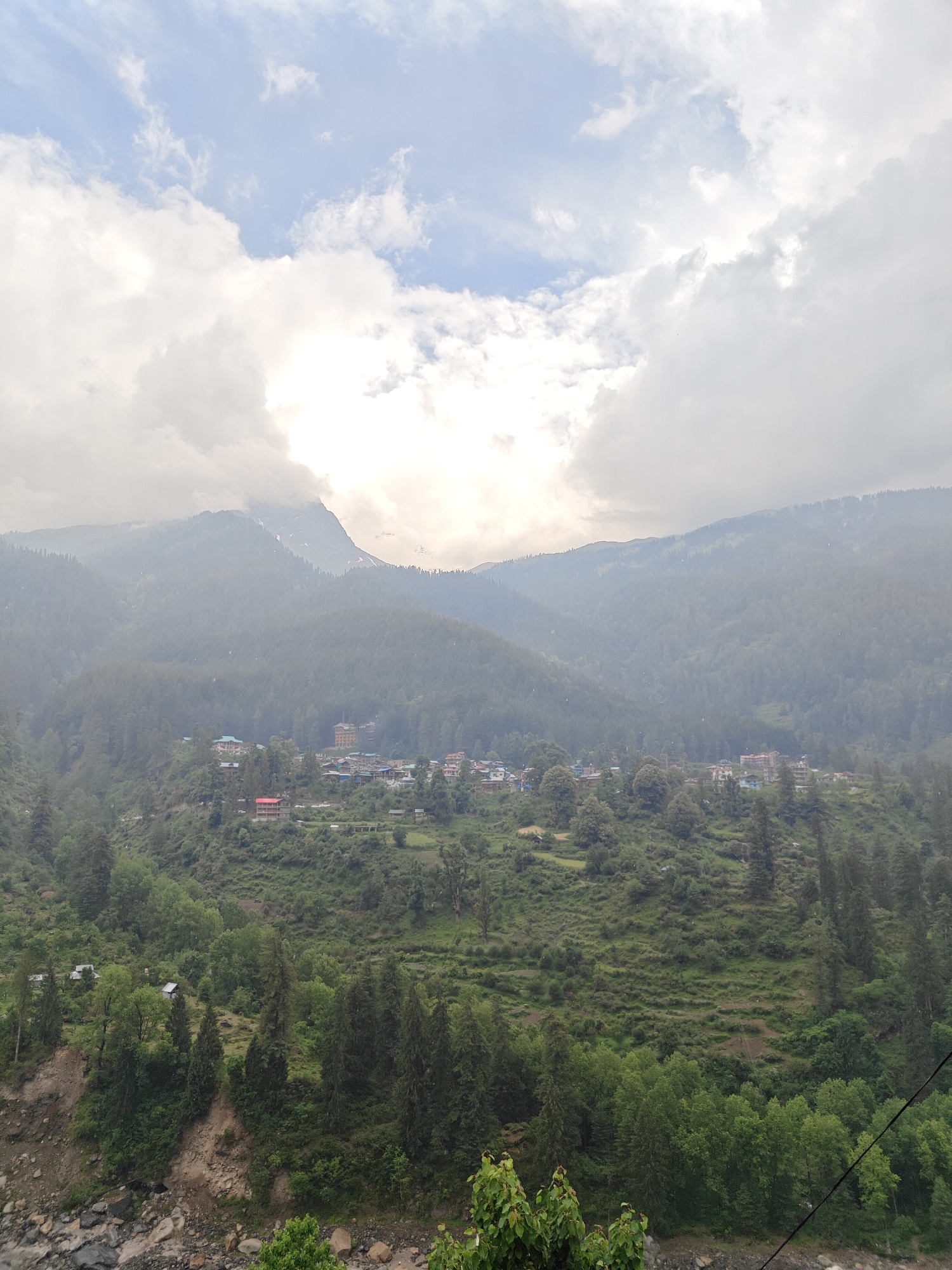 The image presents a wide, elevated view of a scenic mountain valley, revealing a village nestled within a dense forested landscape, characteristic of Pulga, India. The day appears overcast with diffused light, as heavy white and grey clouds dominate the sky, partially obscuring the higher mountain peaks in the background. Patches of blue sky are visible in some areas.
In the foreground, the bottom left shows a rocky riverbed or stream, bordered by steep, earthy banks covered with a mix of rocks and green foliage. Tall, dark green coniferous trees and lighter green deciduous trees densely cover the lower slopes and valley floor. A thin black line, possibly a power cable, stretches across the bottom right.
The midground is dominated by a clustered village, built on terraced slopes. Numerous buildings with varied roof colors, including red and blue, are visible, suggesting a mix of residential structures and possibly guesthouses. These structures are interspersed among trees and green patches, indicating a blend of human settlement and natural environment. Faint roads or paths can be seen winding through the village.
Further back, successive ranges of mountains rise steeply, covered in thick, dark green forests. The distant peaks are veiled in a soft haze and heavy cloud cover, with glimpses of lighter-colored terrain, possibly snow or rock, peeking through. The scene conveys a sense of tranquility and remoteness, showcasing the symbiotic relationship between human life and the rugged, verdant Himalayan landscape. No people or visible text are present in the image.