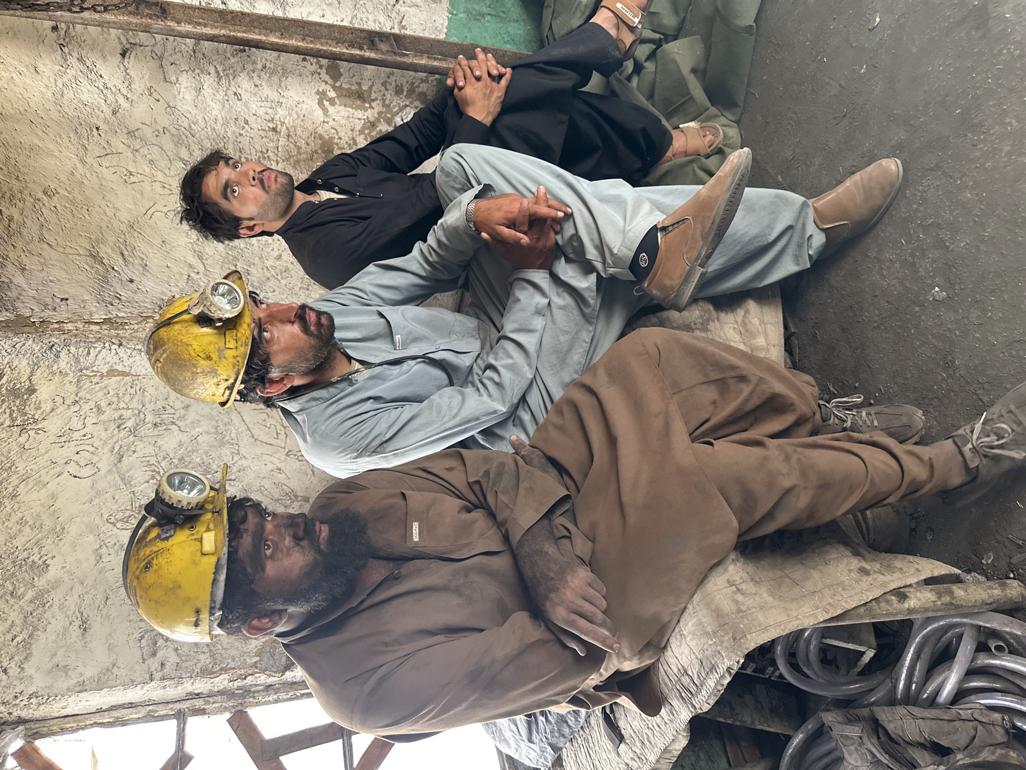 Three men are seated on a raised wooden bench in what appears to be a workshop or storage area in Quetta, Pakistan. The man on the left is wearing a dirty yellow hard hat with a headlamp, a brown tunic, and has a dark beard and face smudged with dirt. He is looking to his right. The man in the middle is wearing a light blue tunic and pants, also with a yellow hard hat and headlamp. He is looking upwards. The man on the right is wearing a black tunic and pants and has his hands clasped in his lap. He is looking to his left. The environment is industrial, with concrete walls, some scaffolding, and coiled pipes visible in the foreground. The lighting is dim, suggesting an indoor, possibly subterranean, setting.