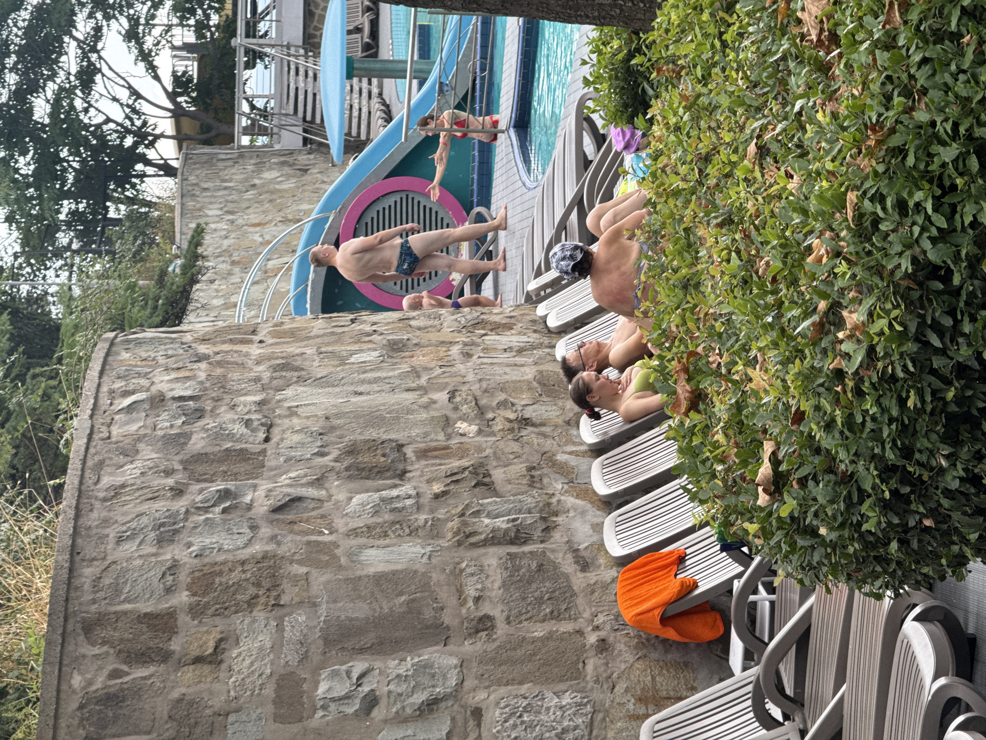 The image depicts a group of people relaxing at a poolside resort. The setting appears to be a sunny day, with clear water in a blue swimming pool and several lounge chairs arranged on a stone patio.
In the foreground, a person is lying on a lounge chair, partially obscured by lush green foliage. Another person is visible on a lounge chair behind them, wearing a green bikini and looking relaxed. Further down the row of lounge chairs, a person with a patterned cap is lying down, and another individual's arm and shoulder are visible. An orange towel is draped over one of the lounge chairs.
In the background, a man in patterned swim trunks is standing near the edge of the pool, and another person, possibly a child, is sitting on the stone wall. A woman in a red bikini is in the swimming pool, reaching out as if to grab onto a railing. A large blue slide structure is visible behind her. The architecture of the resort includes a stone wall and buildings with windows.
The overall mood is one of leisure and relaxation, typical of a summer vacation at a resort. The scene is brightly lit, suggesting it is daytime. There is no visible text in the image.