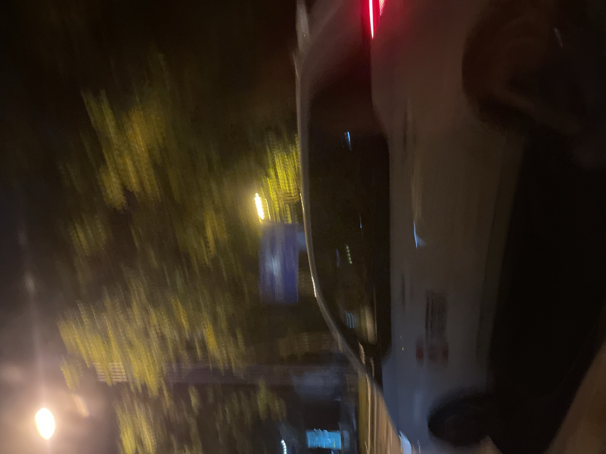The image captures a blurry nighttime street scene in Panyu District, China. A white vehicle is prominent in the foreground, with its side and rear visible. The vehicle appears to have some markings on its side, which are indistinct due to motion blur. The rear taillight of the car is illuminated red. In the background, streetlights cast a yellow glow on blurred trees and foliage. A street sign with blue and white elements is also visible, along with some illuminated vertical blue signage on the left. The overall impression is one of movement, likely captured from another moving vehicle.