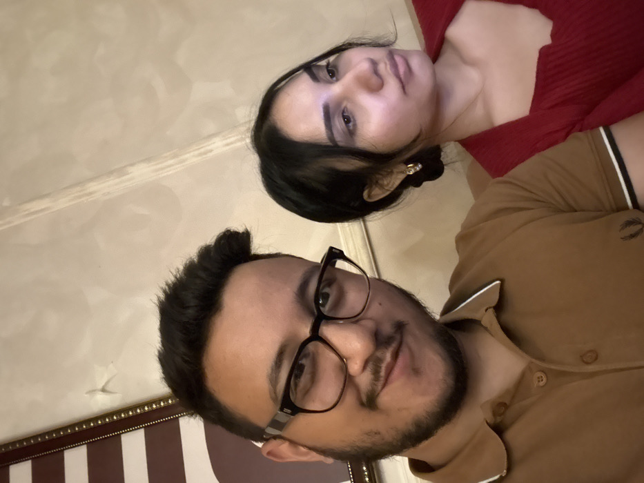 A man with dark hair and glasses and a woman with dark hair are in the picture. The man has a neatly trimmed beard and is wearing a brown polo shirt. The woman is wearing a red top and a small earring is visible. The background is a beige wall with a decorative trim. A portion of a framed print with vertical stripes is visible at the bottom. The angle of the shot is tilted.