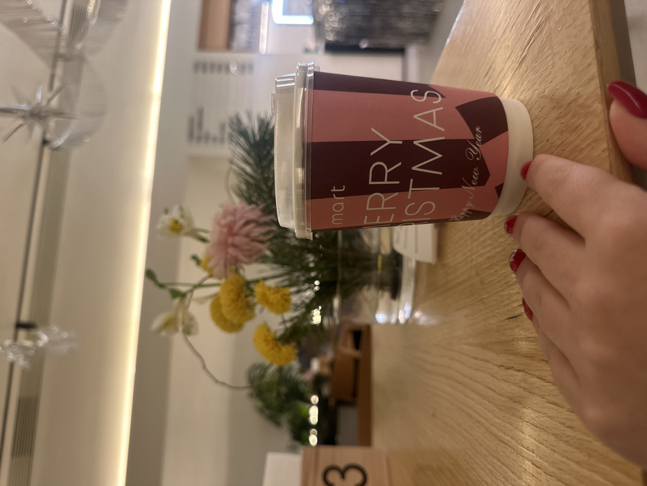 A "Merry Christmas" paper coffee cup with a lid sits on a wooden table beside a small bouquet of yellow and white flowers in a clear glass vase. The cup is pink and dark brown. A person's hand with red fingernails is partially visible near the cup. A blurred interior scene with a light fixture and shelving is in the background. The table has a light wood grain.